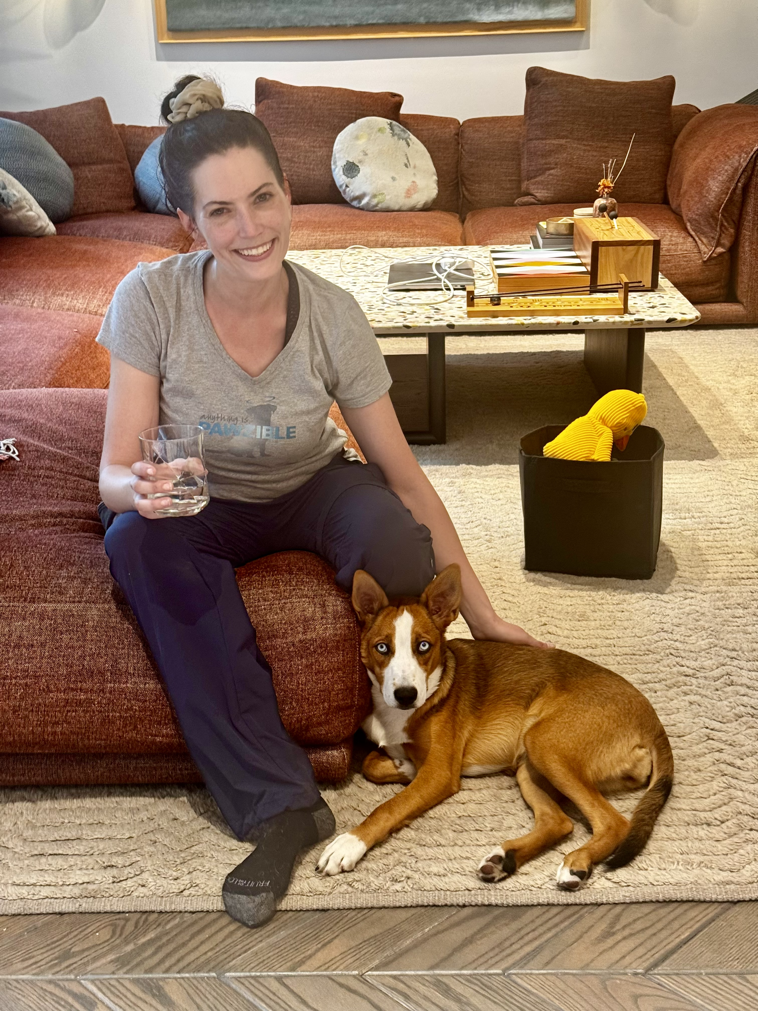 A woman sitting on a red sofa with a glass of water in her hand, smiling. A brown dog with blue eyes lying next to her. The living room has a coffee table with books and decorative items, and a black storage bin with yellow slippers inside.