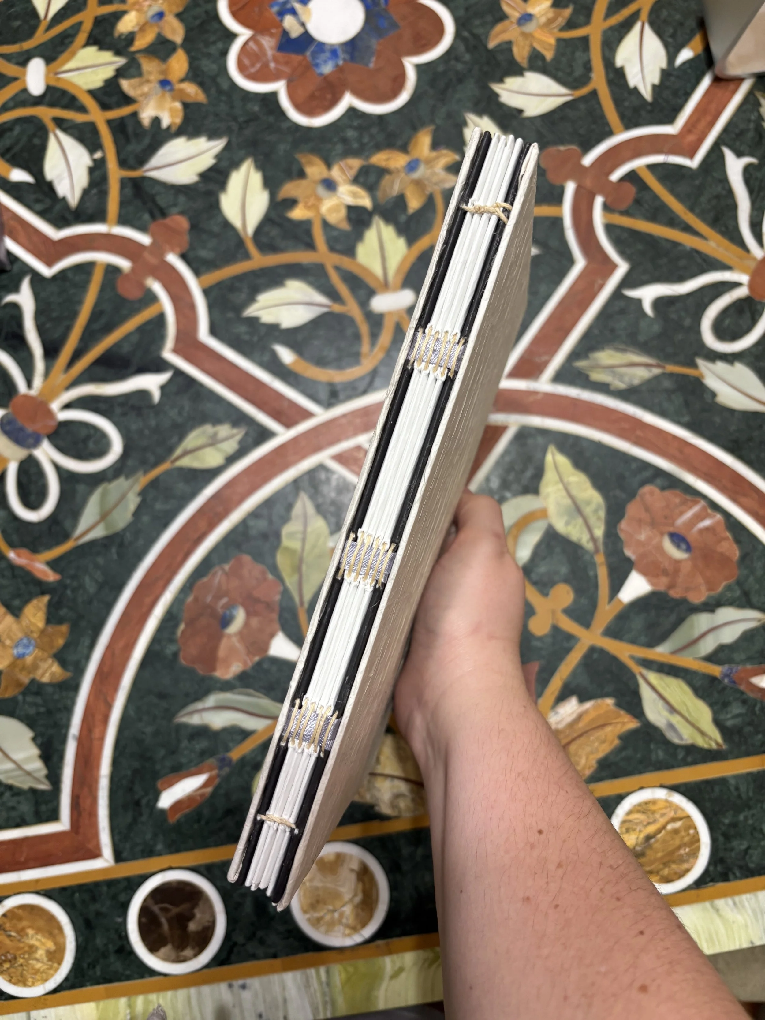 A person's hand holding a book or notebook with the spine facing upward, showing the binding, against a colorful floral patterned tile floor.