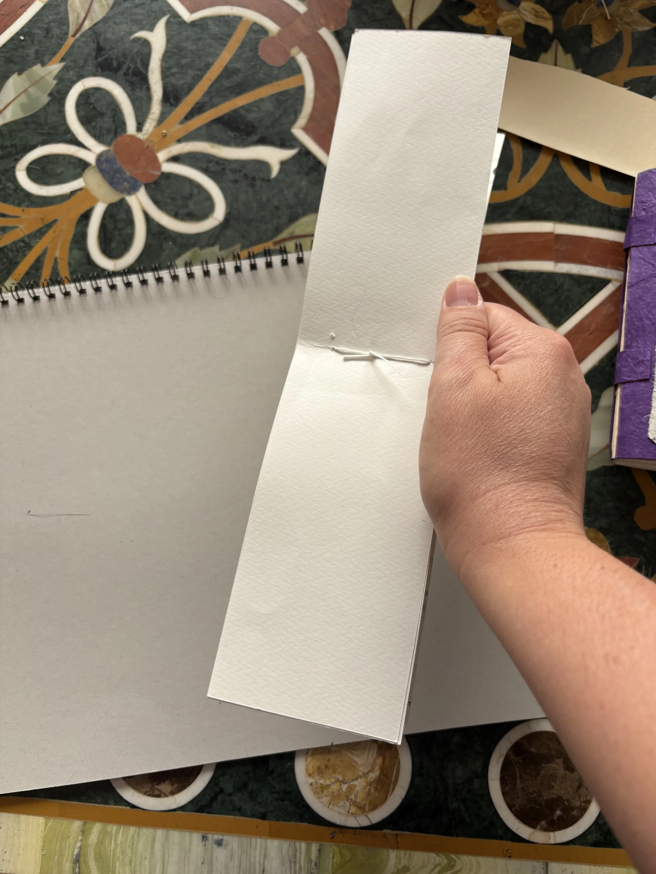 A hand holding open a blank watercolor paper pad with a textured surface, resting on a colorful table with mosaic-like floral patterns.