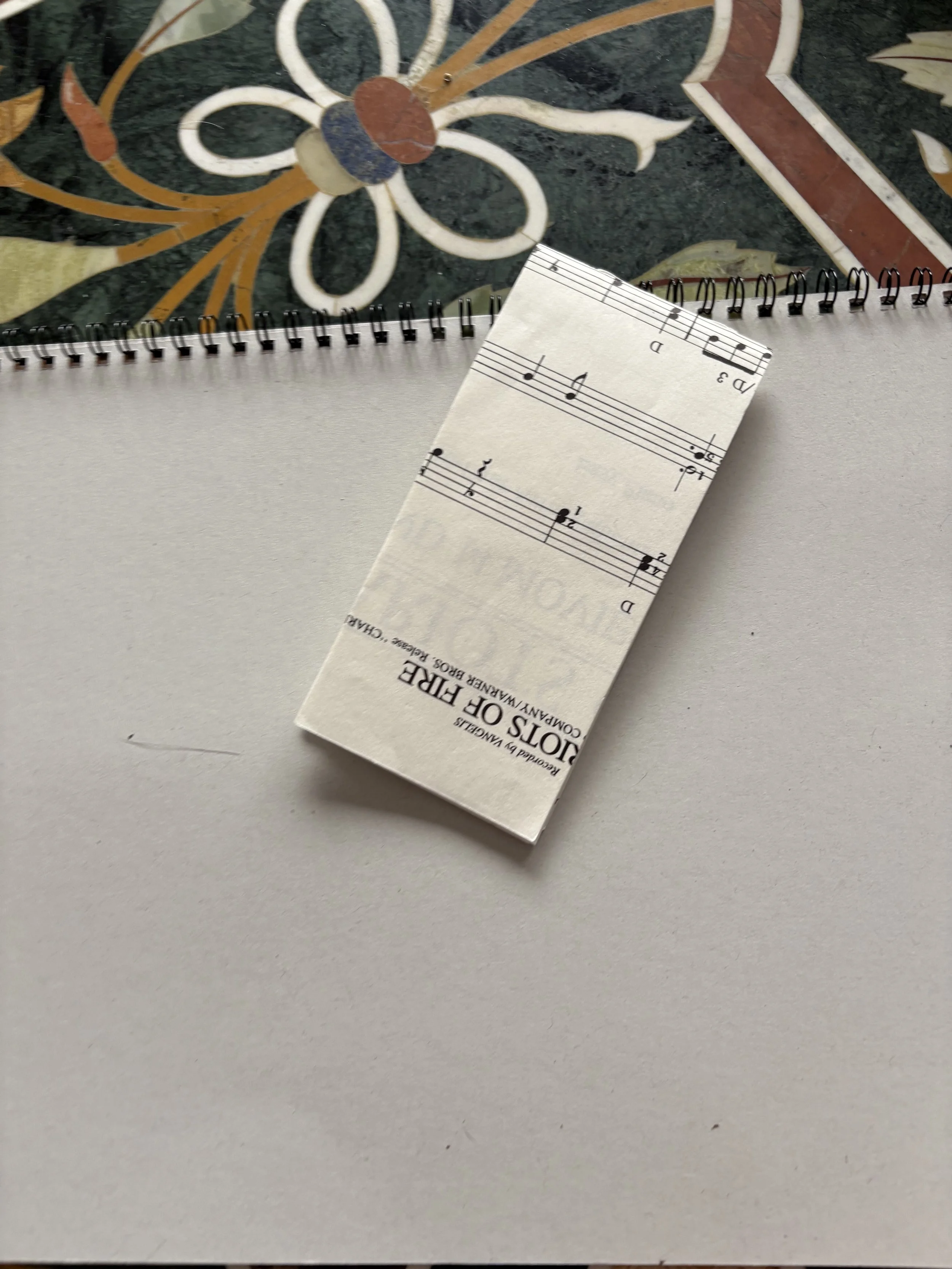 A music score pamphlet labeled 'Voice of Fire' resting on a white surface with a spiral-bound notebook and an ornate tile or mural in the background.