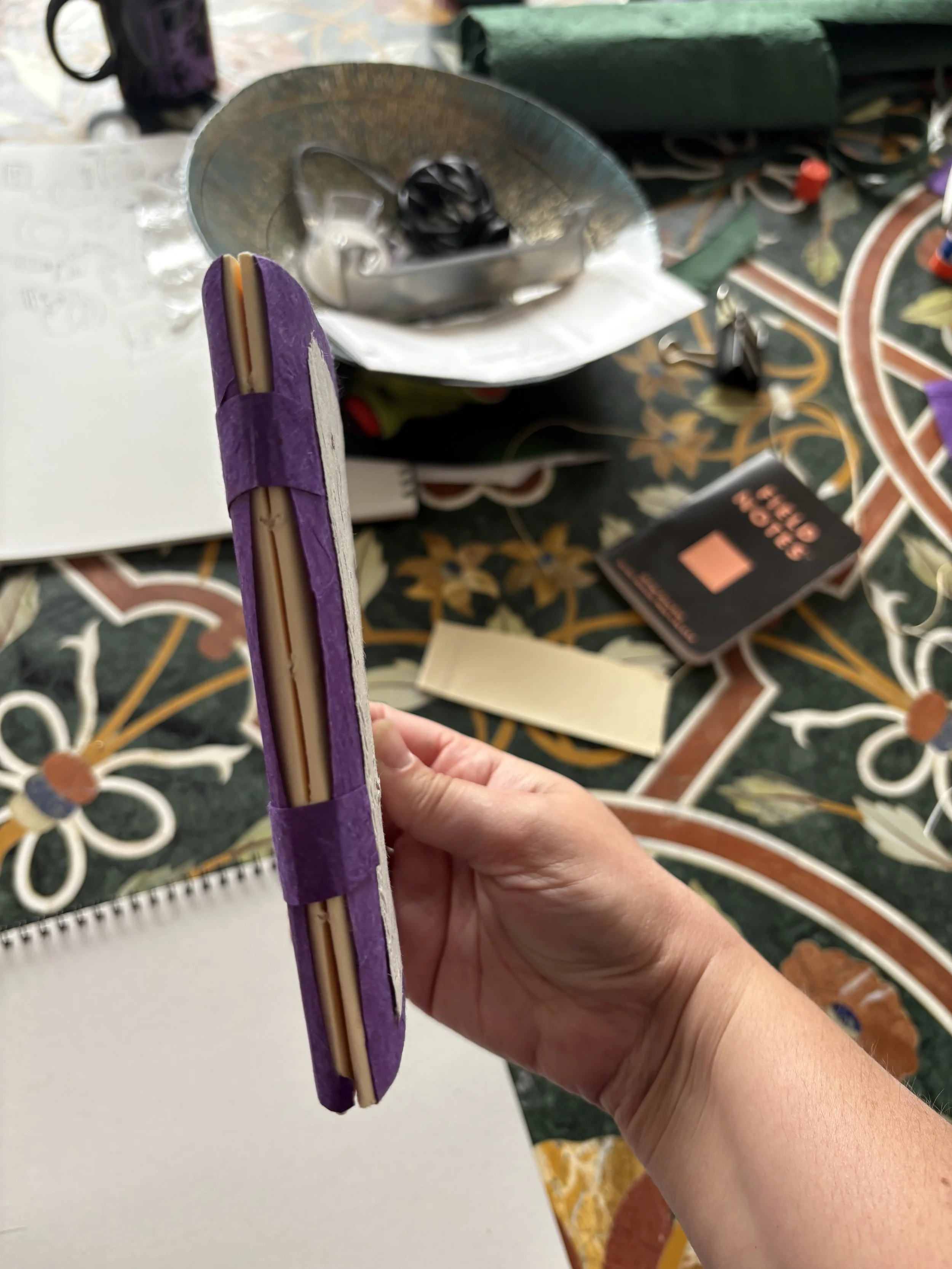 A hand holding a purple and beige folder with papers inside, on a table with various objects including a notepad, a book, a bowl, and miscellaneous items.