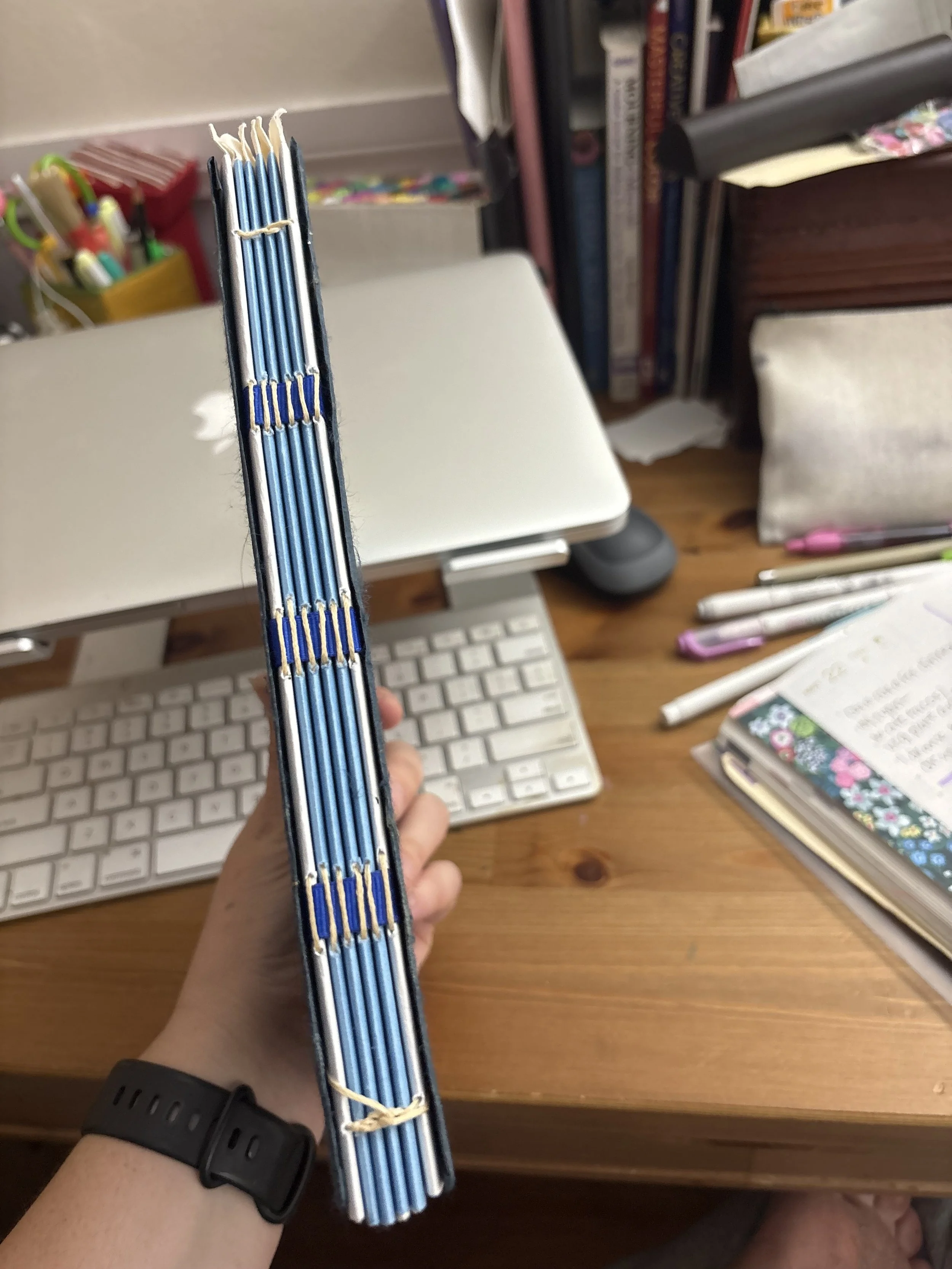 Hand holding a trio of notebooks with blue covers, stacked together over a cluttered desk with a computer keyboard, pens, and books.
