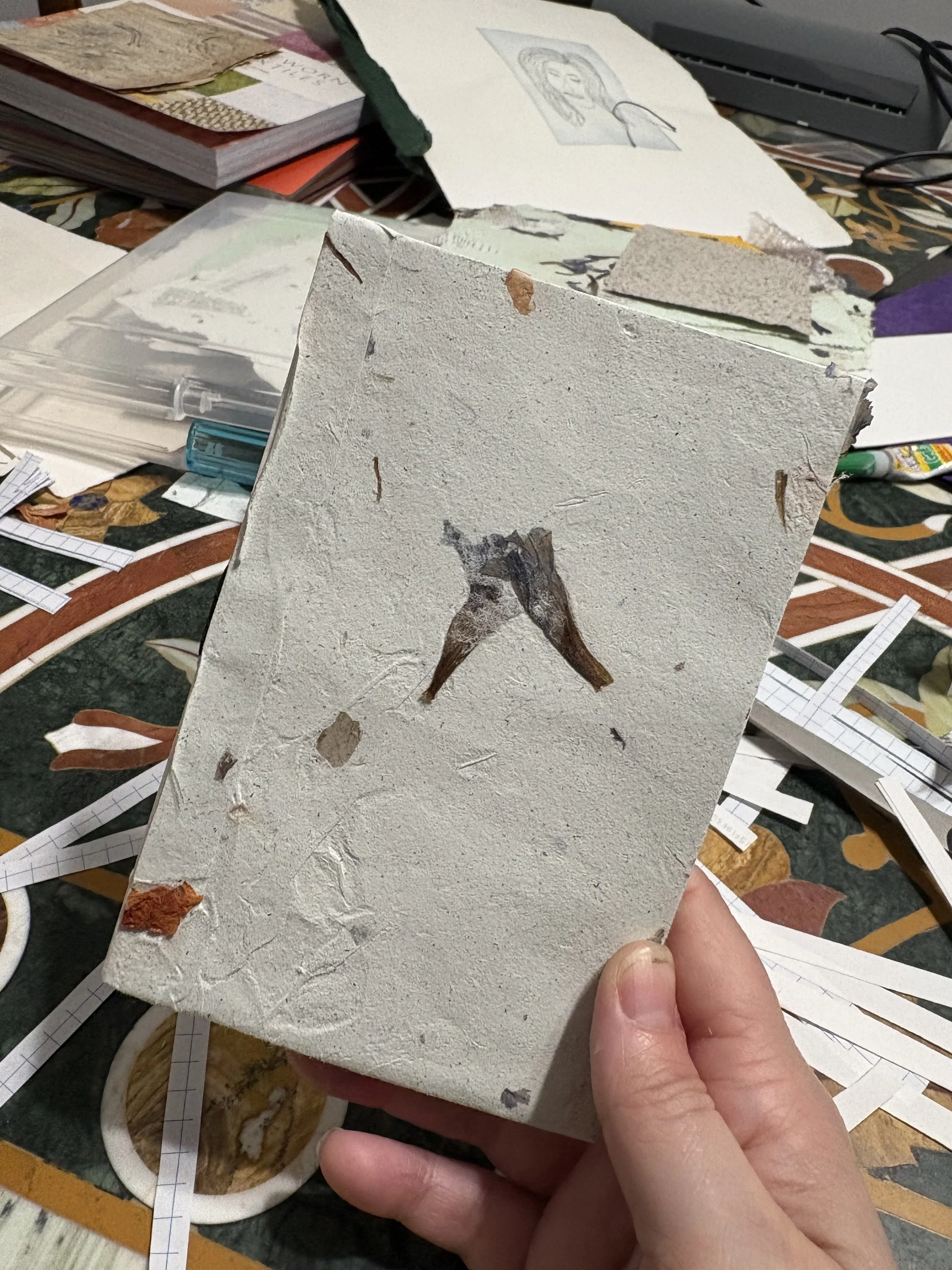 A person holding a textured piece of paper or artwork with a small, painted image of a bird on it. The background is cluttered with various papers, notebooks, and art supplies.