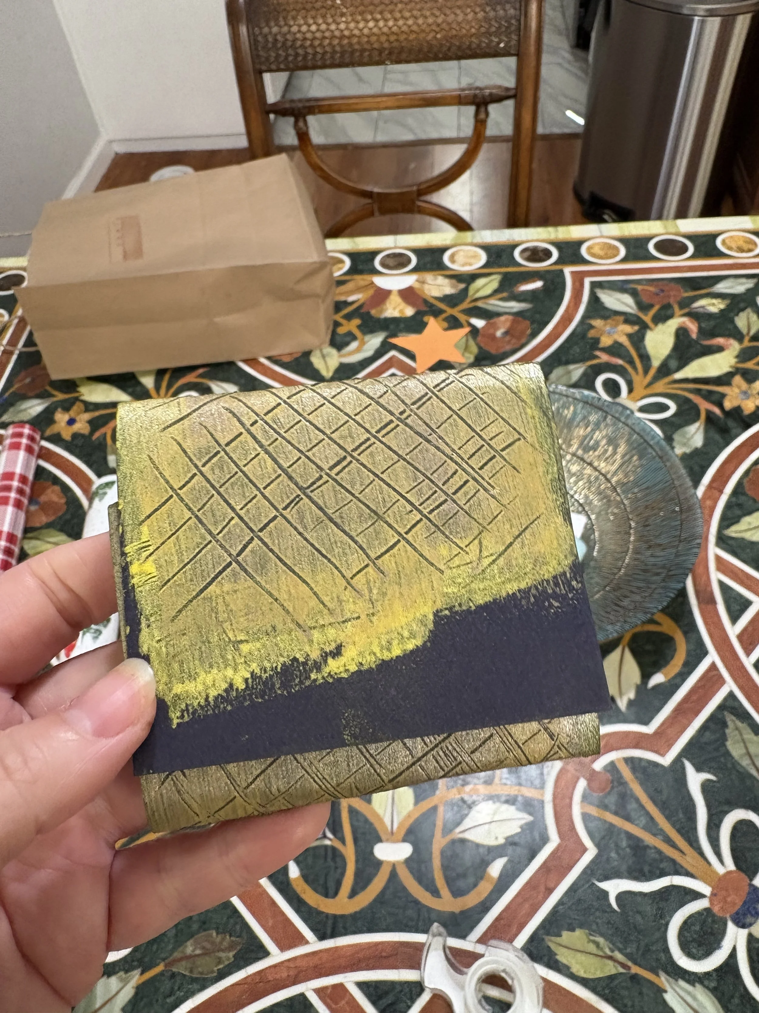 Hand holding a small card with a textured yellow and black design, featuring a pattern of parallel lines cut into the surface, in a room with a patterned tablecloth, a brown paper bag, a glass plate, a paper towel roll, and a wooden chair.