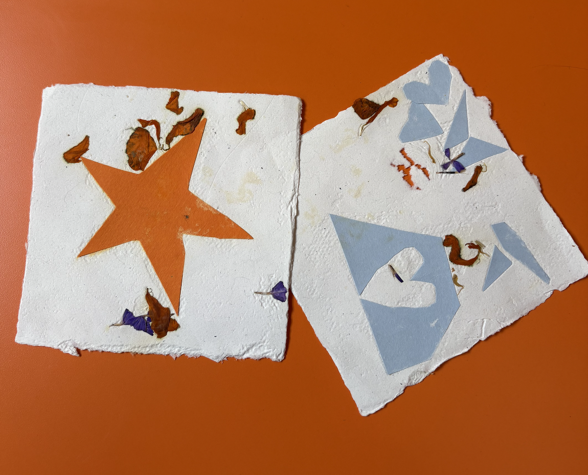 Two handmade paper cards, one with an orange star cutout and the other with a blue heart cutout, on an orange surface. Both cards have dried flower petals and paper butterflies attached.