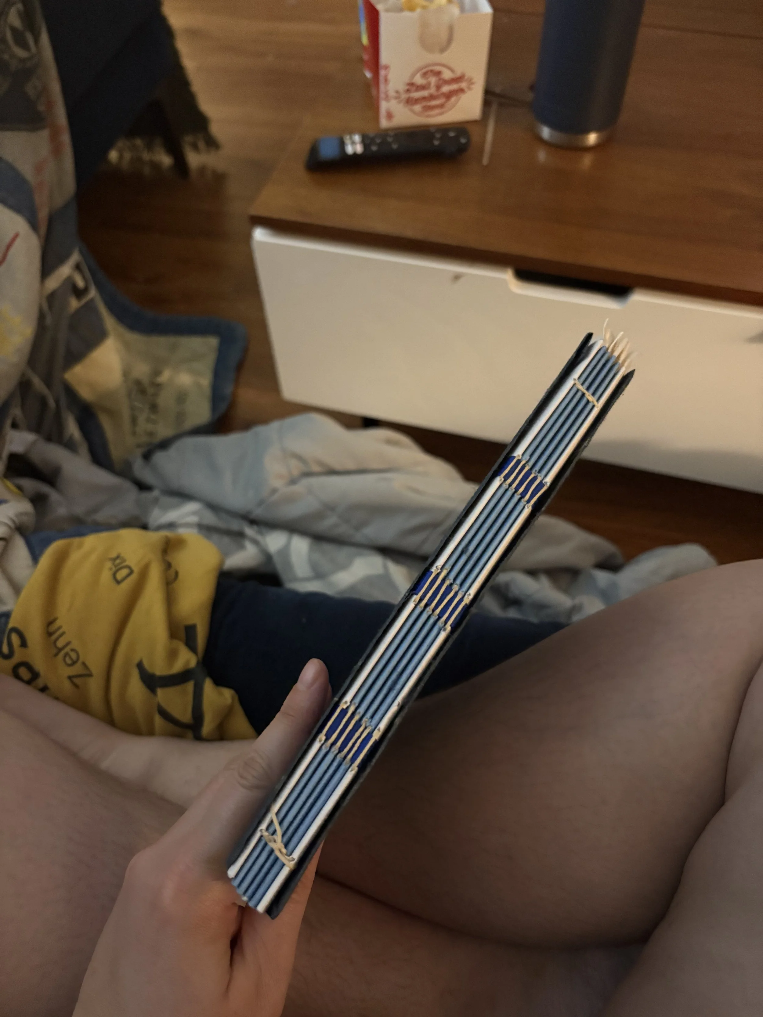 Person sitting on bed holding a book with a blue and white cover, with a nightstand holding a TV remote, popcorn box, and a travel mug in the background.