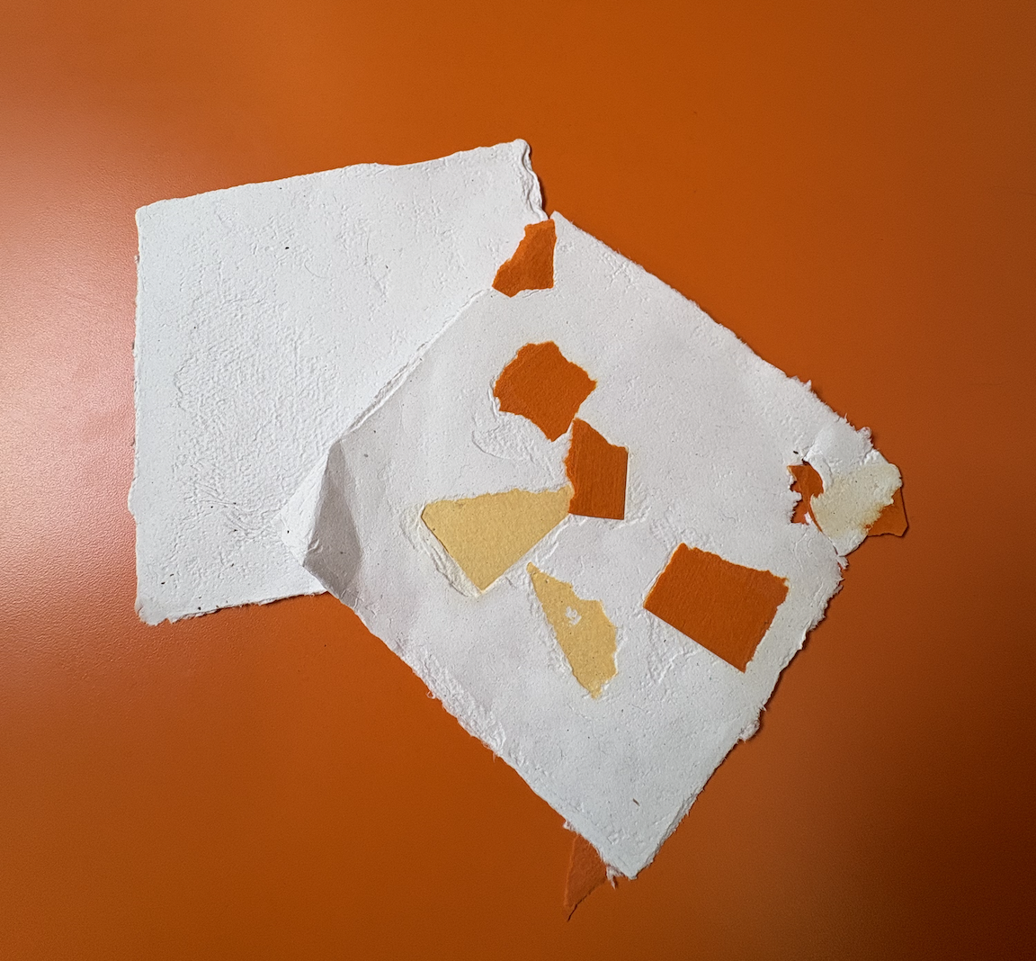 A torn piece of paper with small paper pieces in orange and beige colors on top of a larger, flat, white paper, placed on an orange surface.