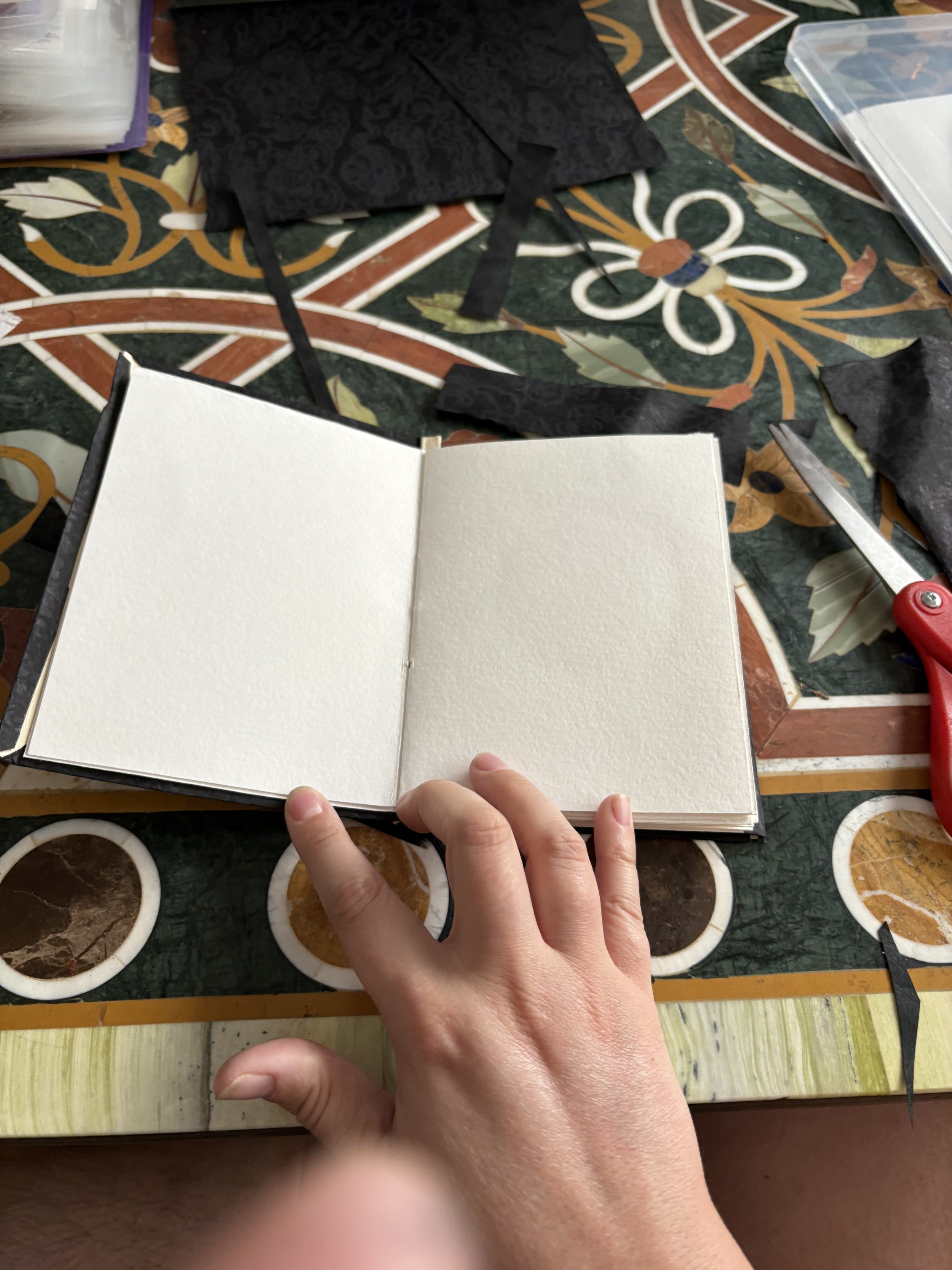 Open sketchbook or photo album on a colorful patterned table with craft supplies, scissors, and black decorative paper scraps around.