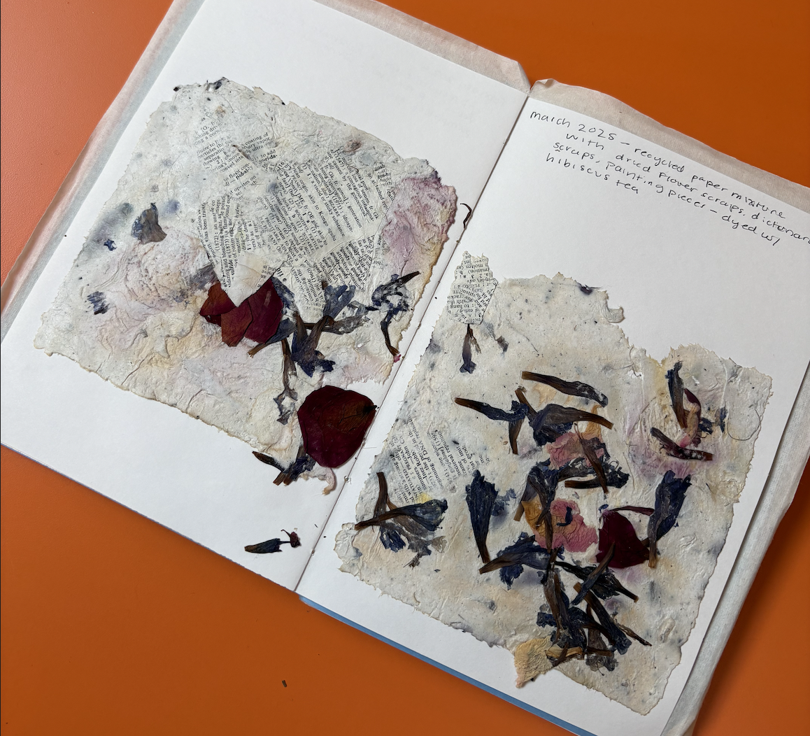 Open scrapbook with collages of torn paper, dried flower petals, and botanical pieces on white pages, placed on an orange surface. Handwritten notes on the right page mention recycled paper, flower scraps, and specific plants.