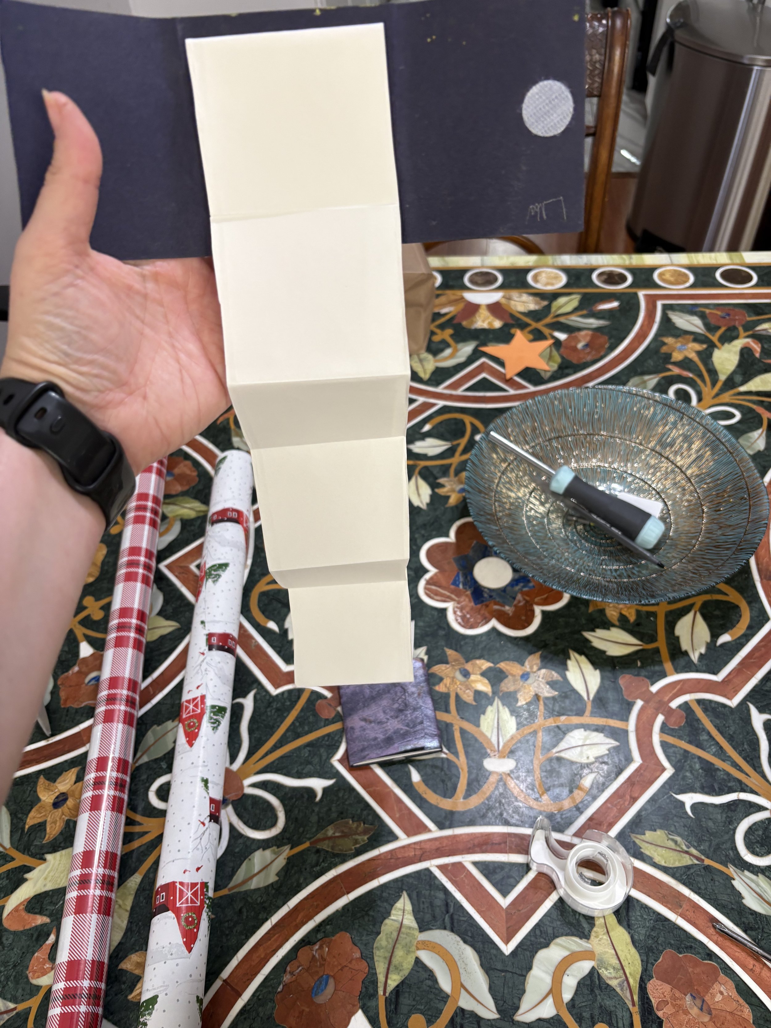 Person holding a black gift box with a white interior, on a table with Christmas-themed wrapping paper, scissors, a pen, a small bowl, and tape.