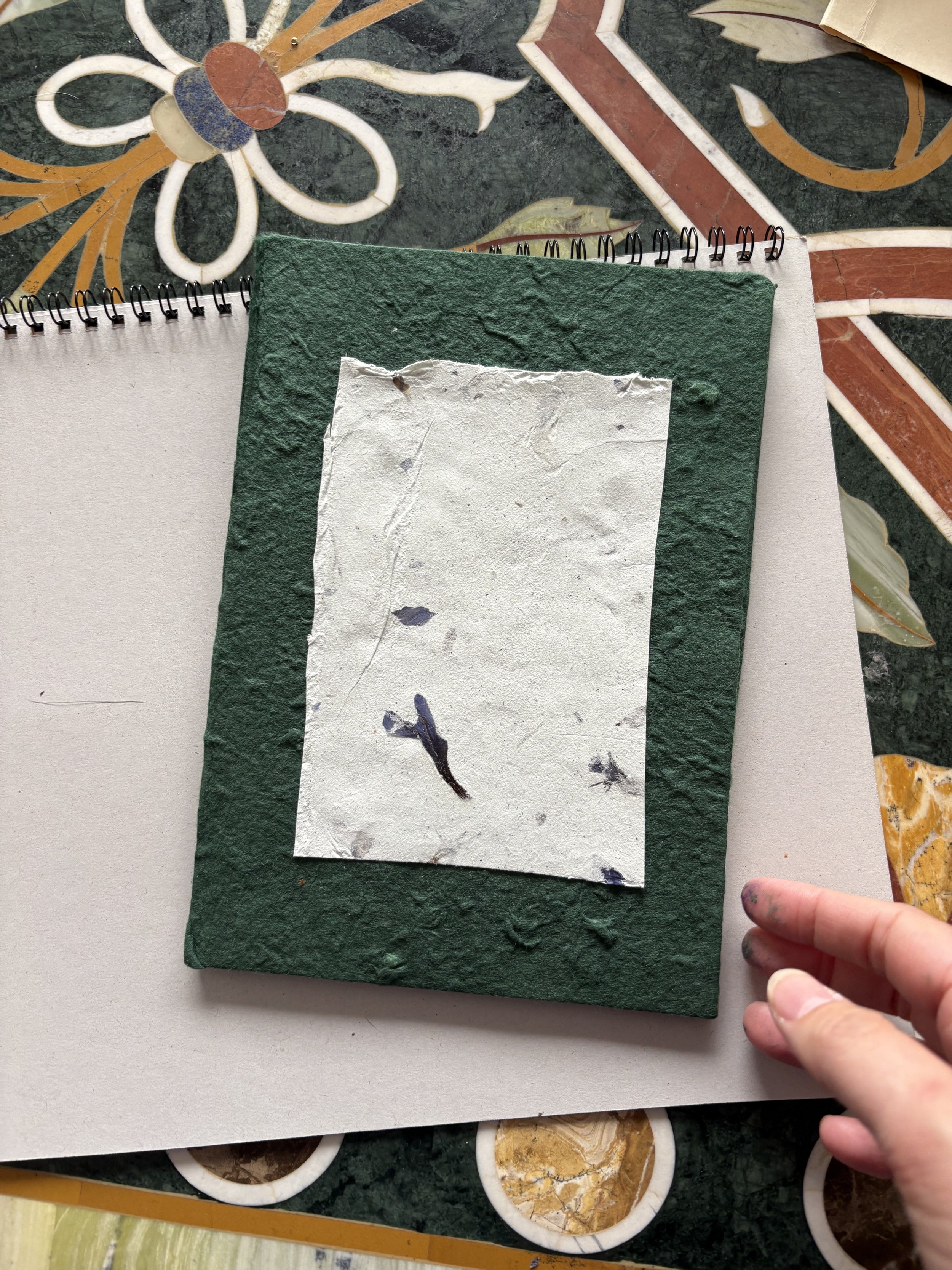 Close-up of a green textured card with a smaller torn white paper glued on top, on a surface with colorful flower and geometric tile patterns, and a person's hand with painted nails is partly visible.