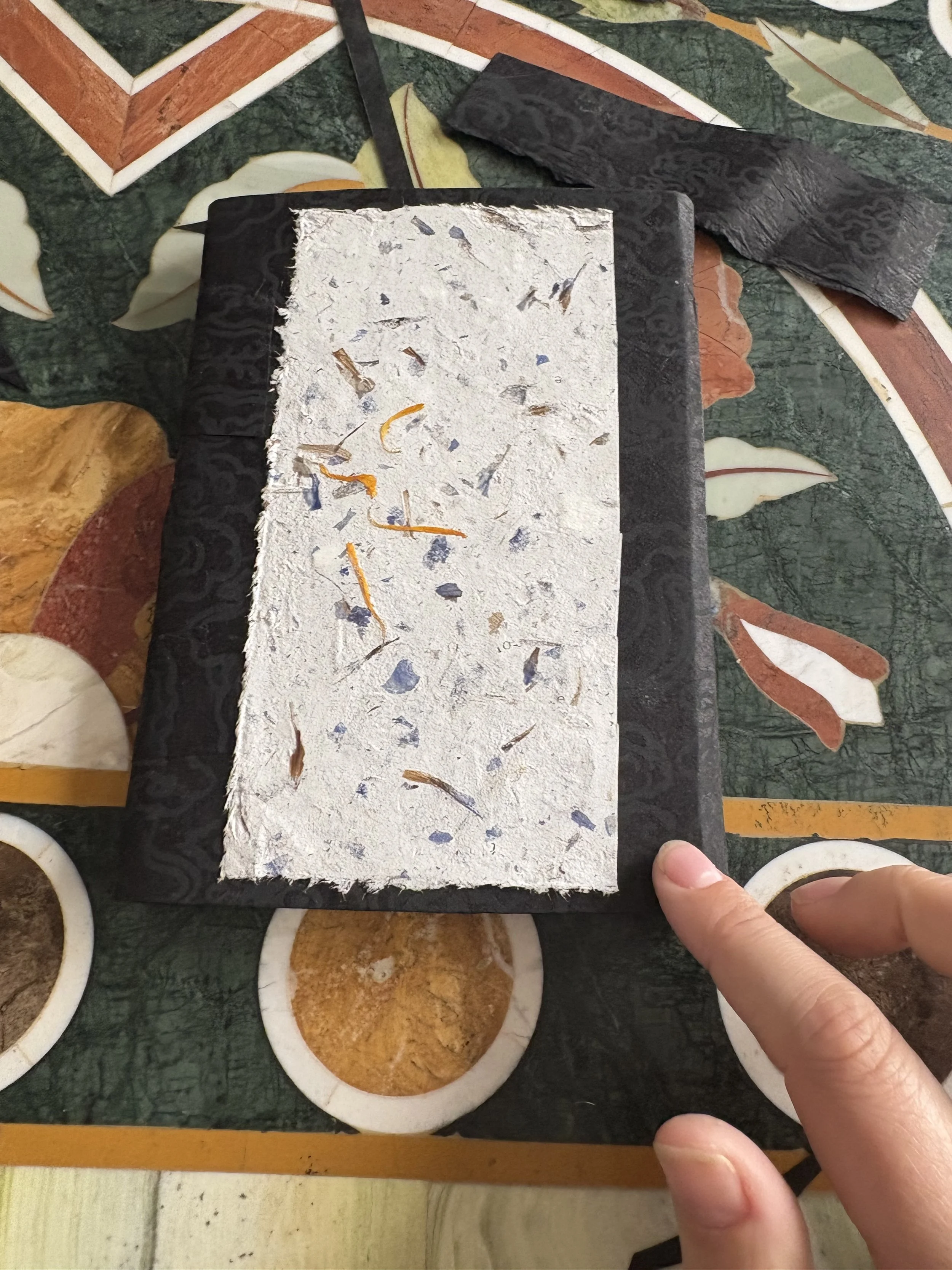 A rectangular piece of paper with colorful shavings on it, placed on a dark, textured surface. A person's hand is visible holding the corner.