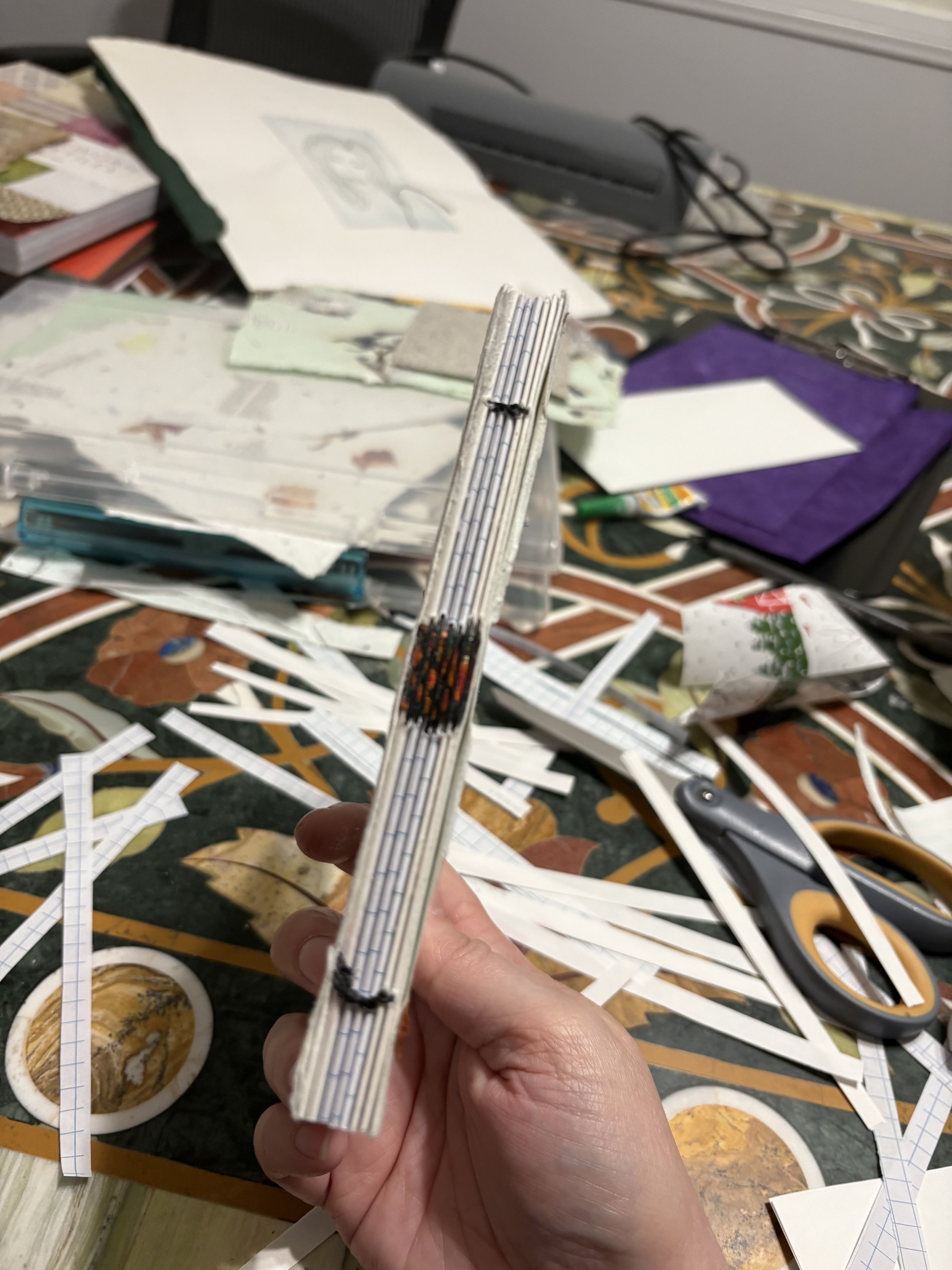 Hand holding a partially constructed paper or cardboard sculpture with a busy workspace in the background.