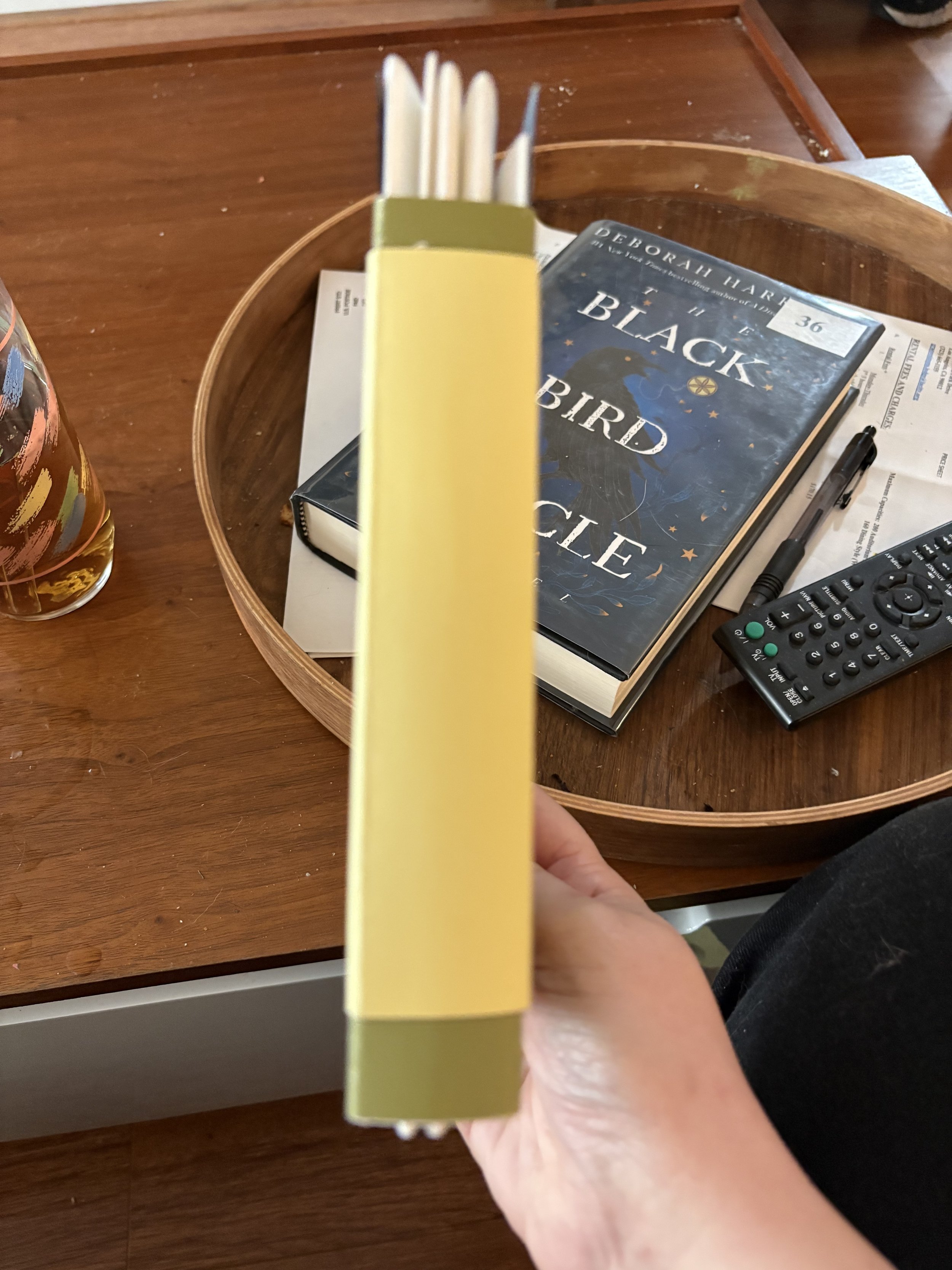 Close-up of the bottom edge of a yellow book held in a person's hand, with the pages visible on top, on a wooden table with a glass of drink, a book titled 'Black Bird' by Deborah Har, a black remote, and a black pen on a paper.