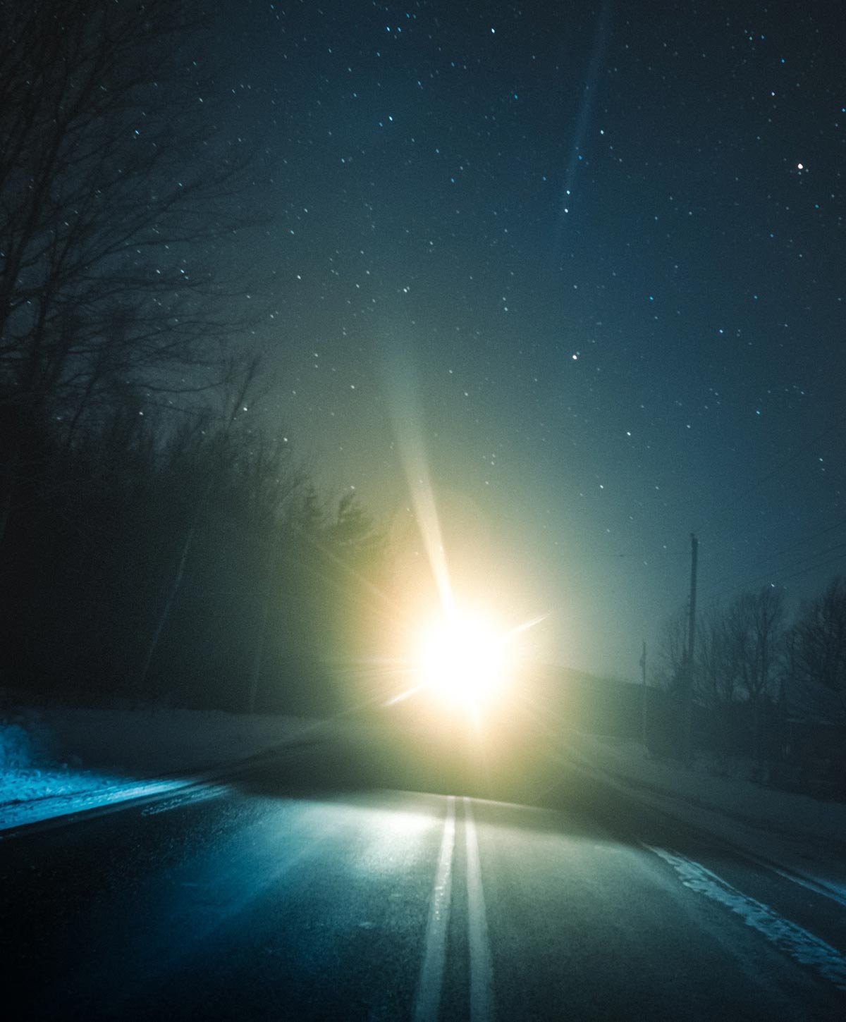 A dark road at night with a bright streetlight or vehicle headlight in the distance illuminating the icy road surface, surrounded by trees and a clear, starry sky.