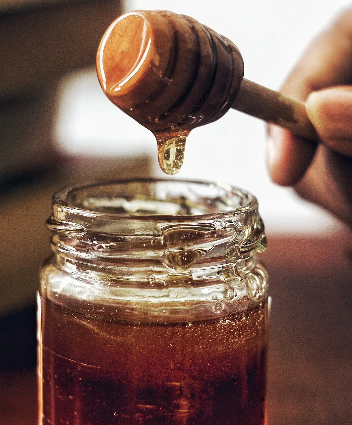 A glass jar filled with honey, with a honey dipper held above it, dripping honey into the jar.