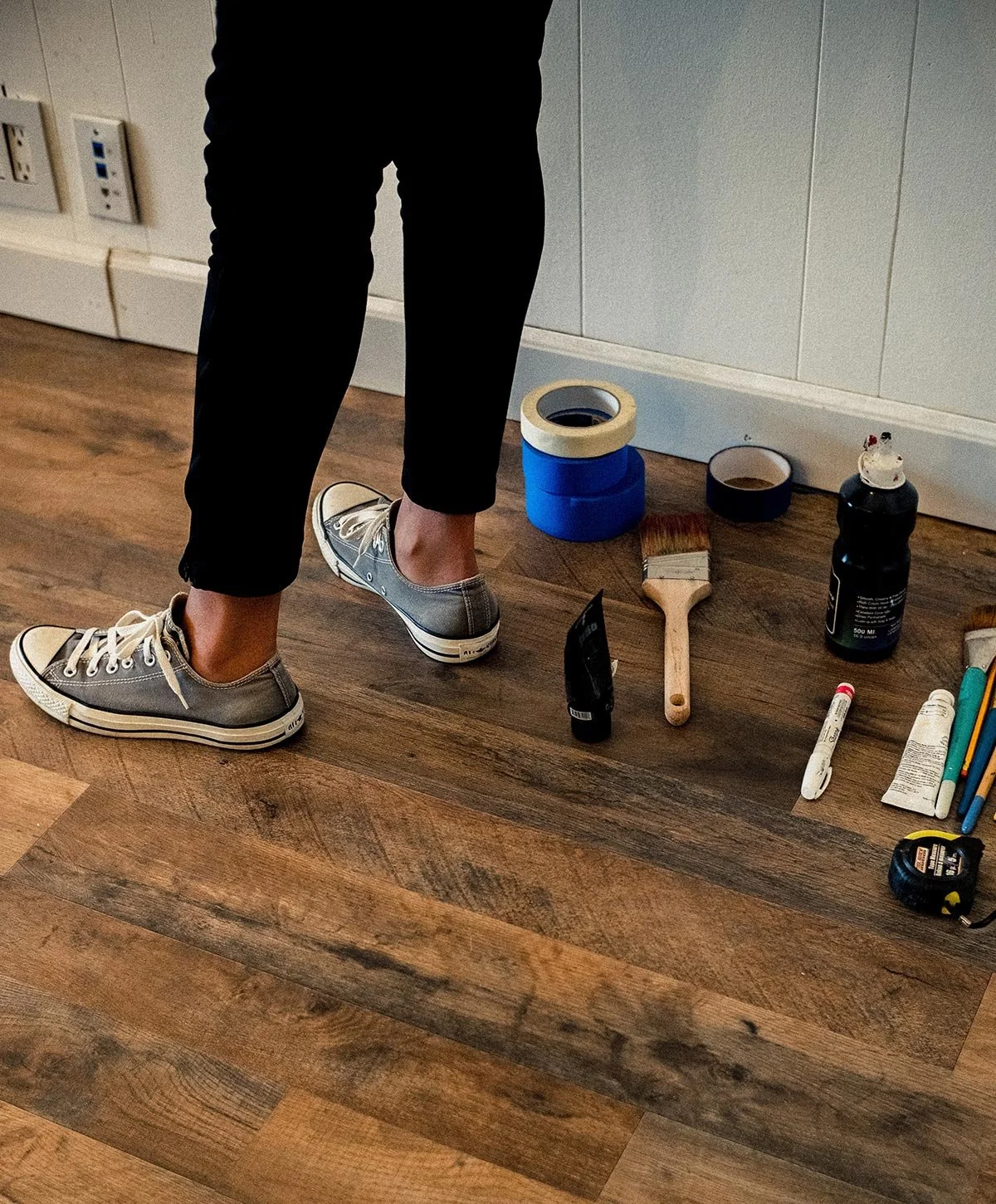 Close-up of a person's legs wearing black pants and gray canvas sneakers, standing on a wooden floor next to various painting tools and supplies, including tape, paint, brushes, and markers.