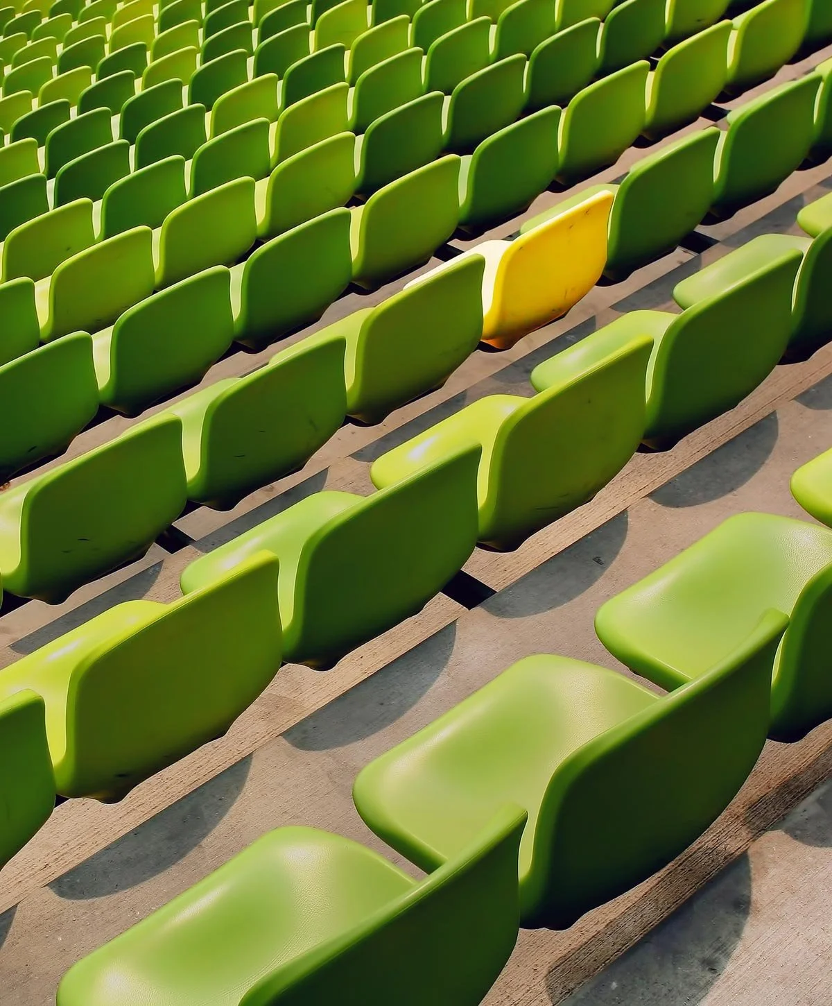 Multiple green stadium seats with a single yellow seat among them, casting shadows on the ground.