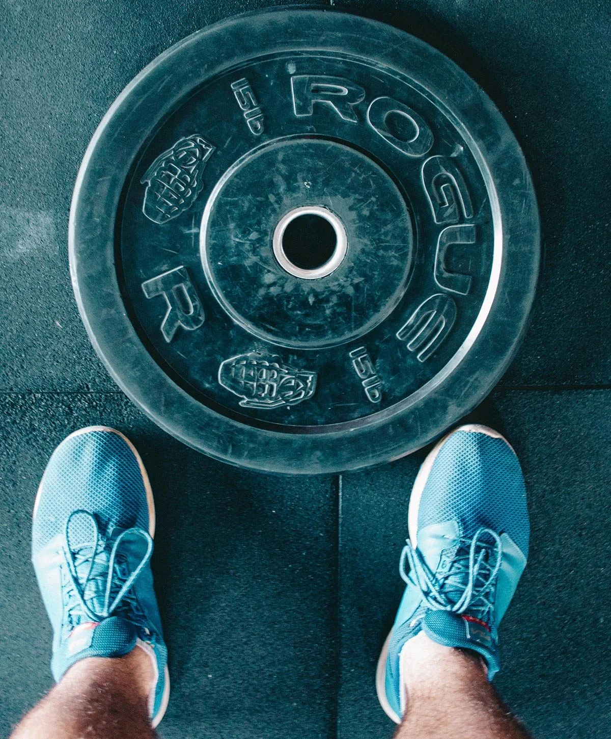 A person wearing blue athletic shoes standing on a rubber gym floor next to a large transparent weight plate with the word 'ROGUE' embossed on it.
