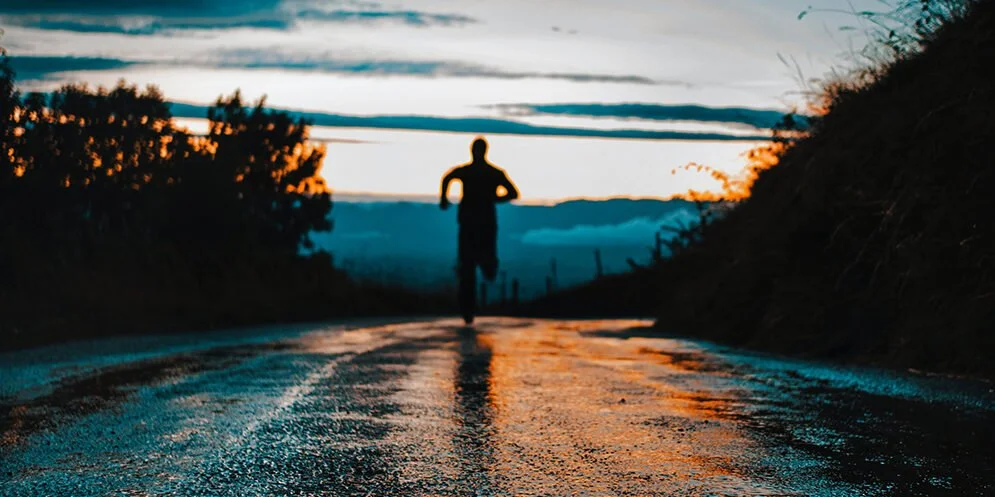 A person running on a wet road during sunset with trees and hills on either side.