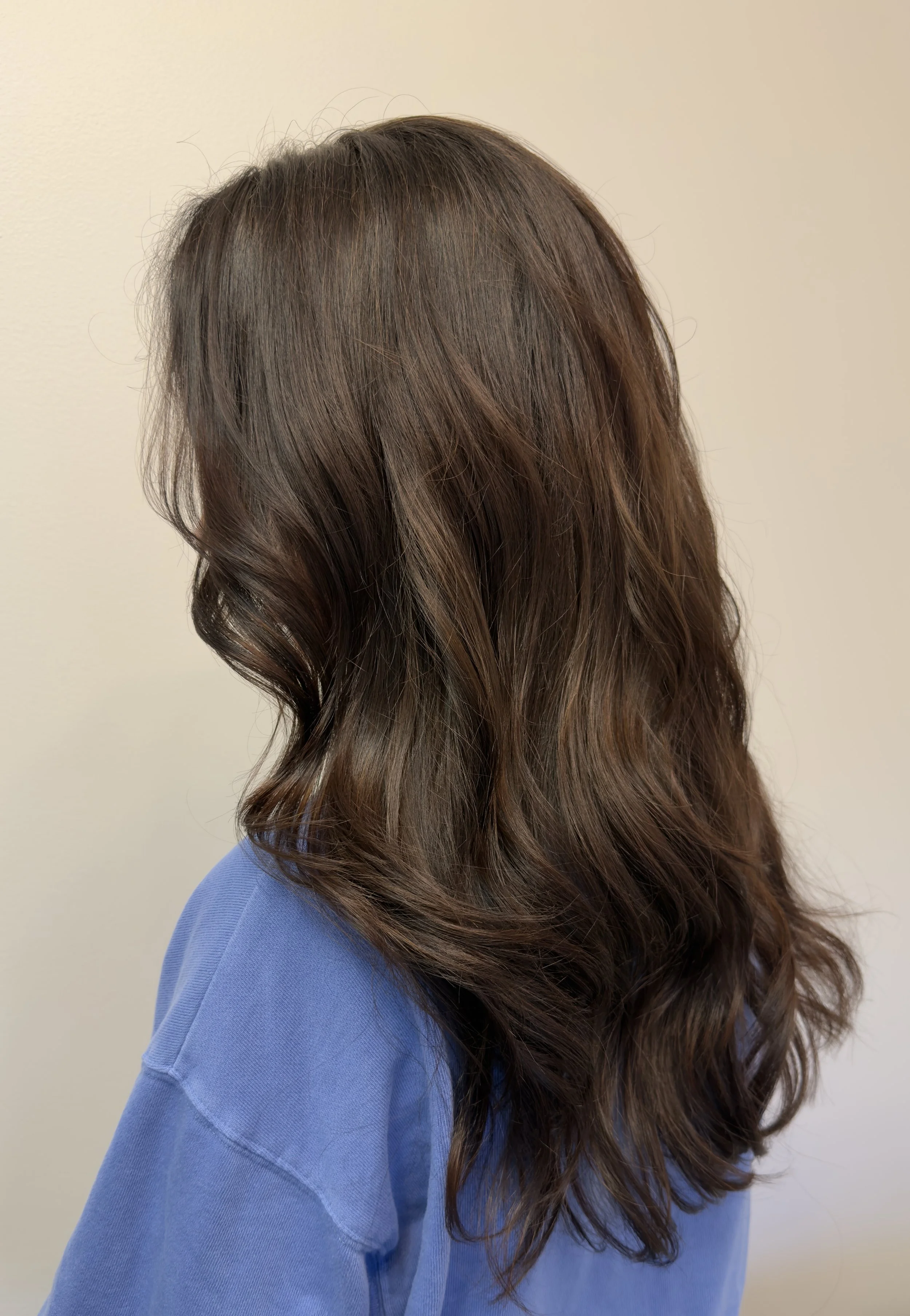 Side view of a woman with wavy, medium-length brown hair wearing a blue shirt, standing against a plain wall.