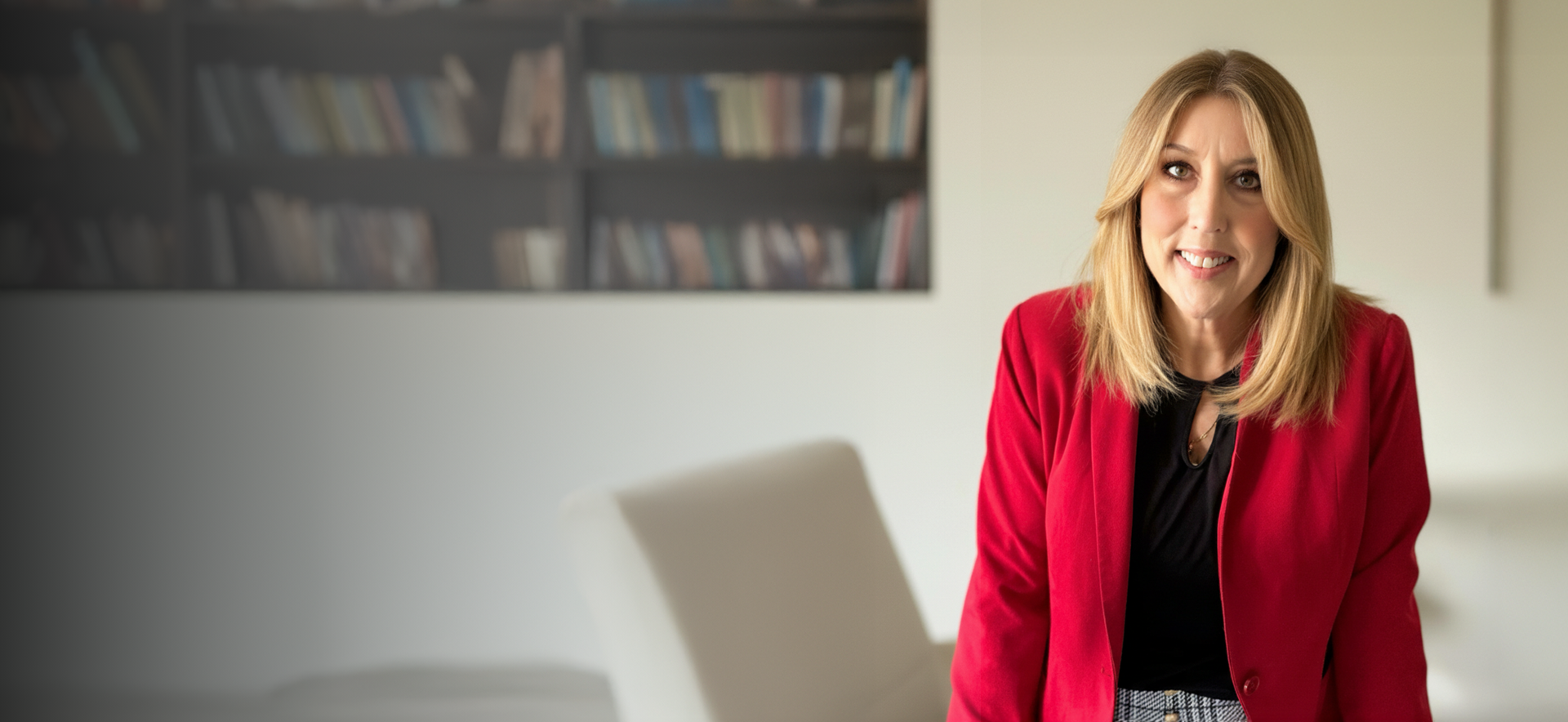 Woman with blonde hair wearing a red blazer and black top, smiling in an office setting.