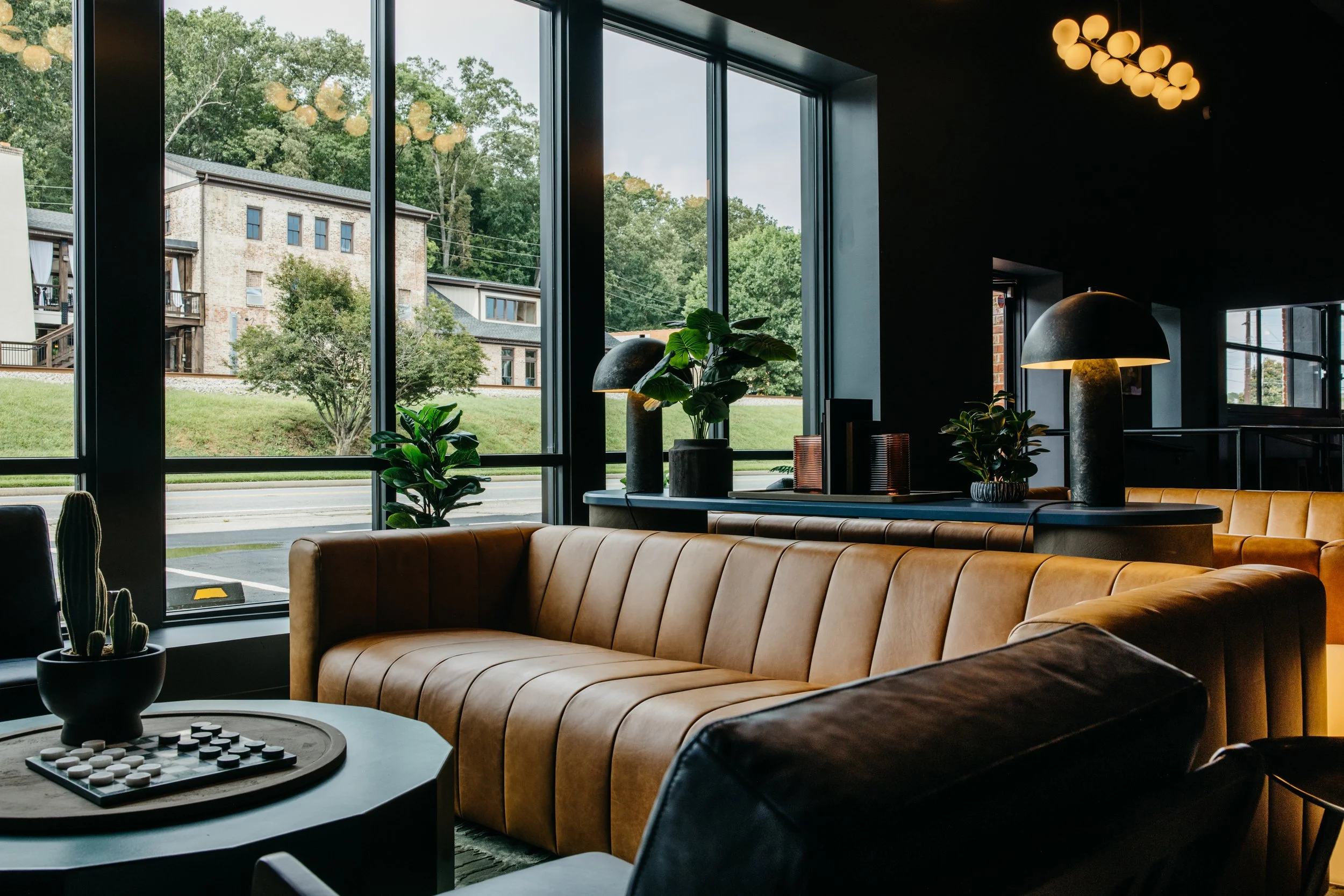 Interior of a modern lounge with large windows, tan leather sofas, potted plants, and decorative lamps, with a view of trees and buildings outside.
