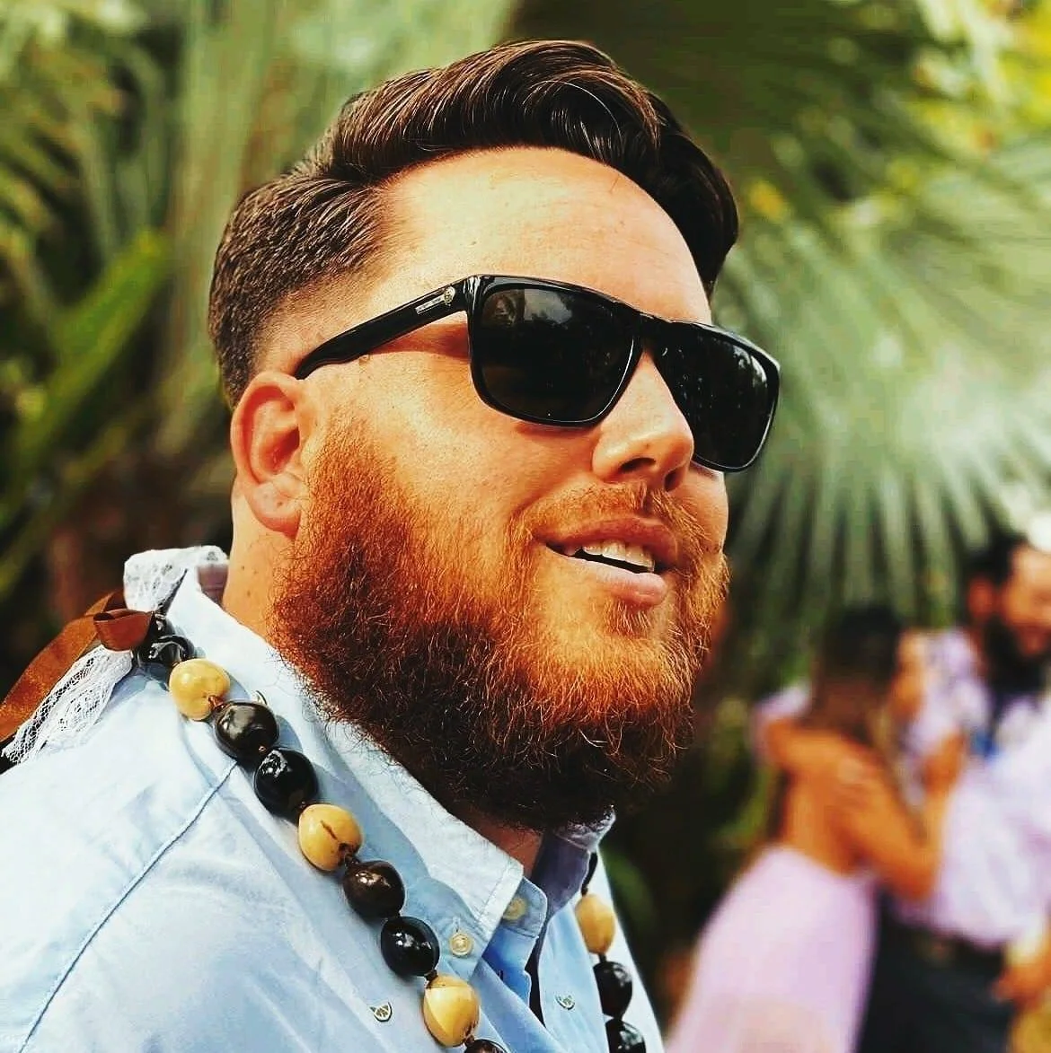 A man with a beard wearing sunglasses, a light blue shirt, and a beaded necklace, outdoors with palm trees and blurred people in the background.