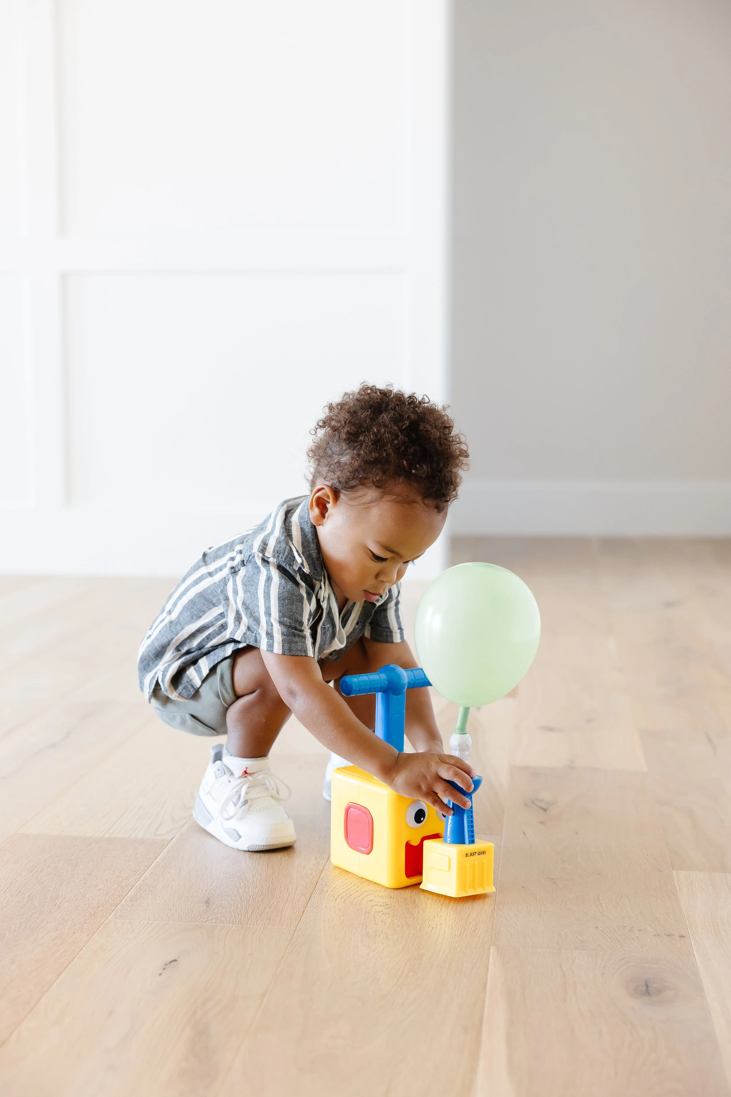 A young child playing with a colorful toy that has a balloon attached, on a wooden floor in a bright room.