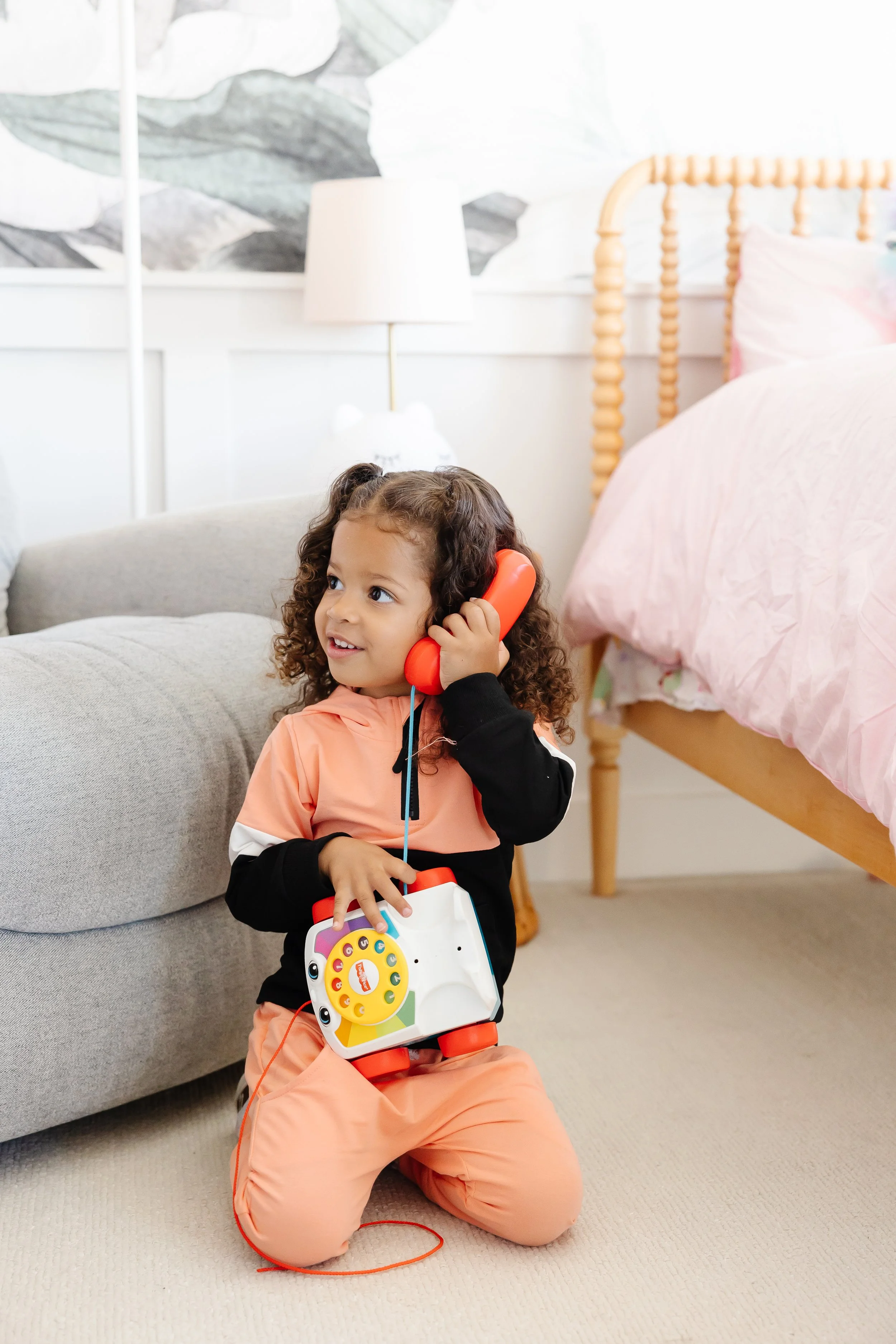 A young girl with curly hair kneeling on a beige carpet, holding a toy telephone with a yellow rotary dial, and talking into a red toy telephone receiver.