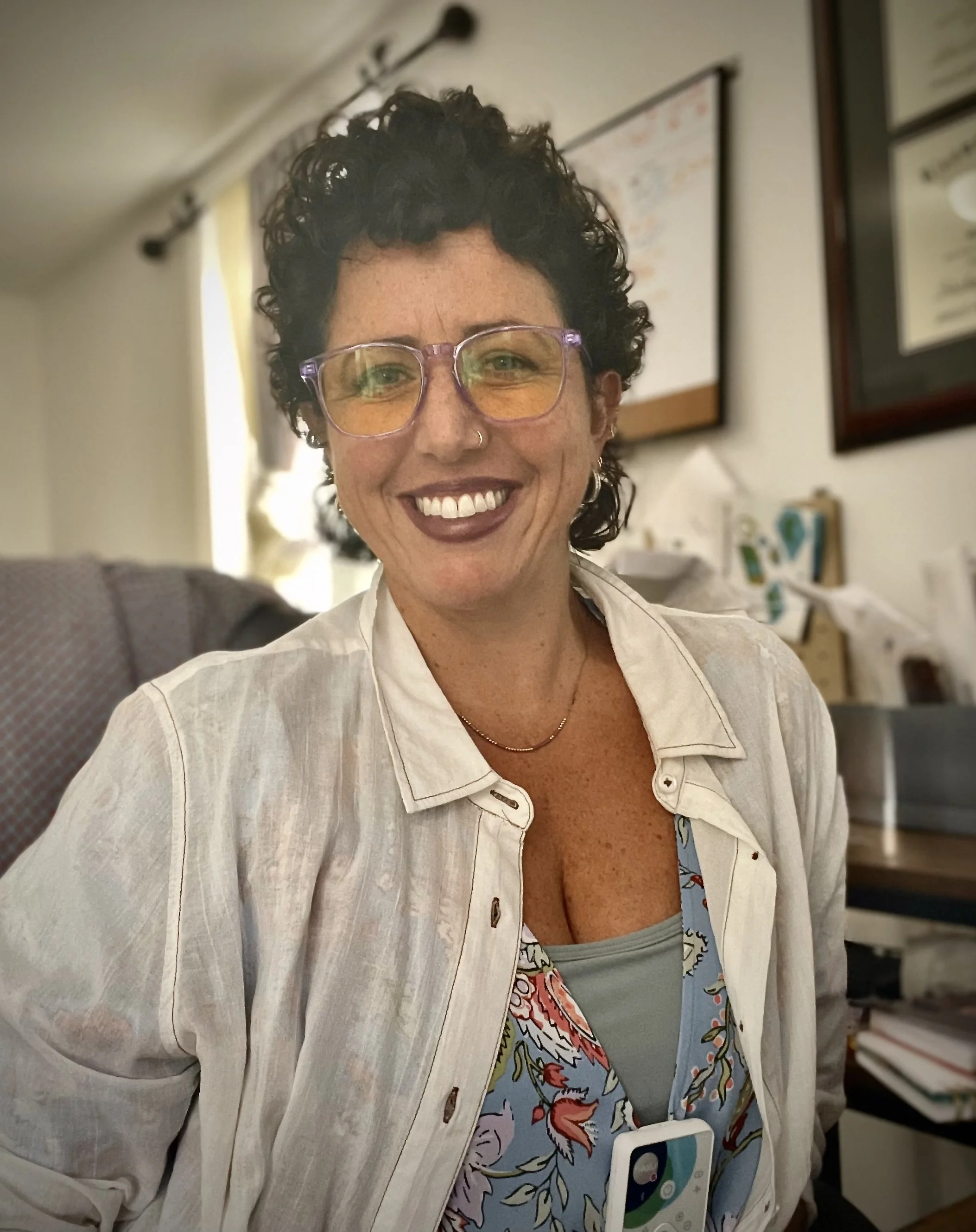 A woman with short, curly dark hair and yellow-tinted glasses smiling in an indoor setting with framed certificates or diplomas on the wall behind her.