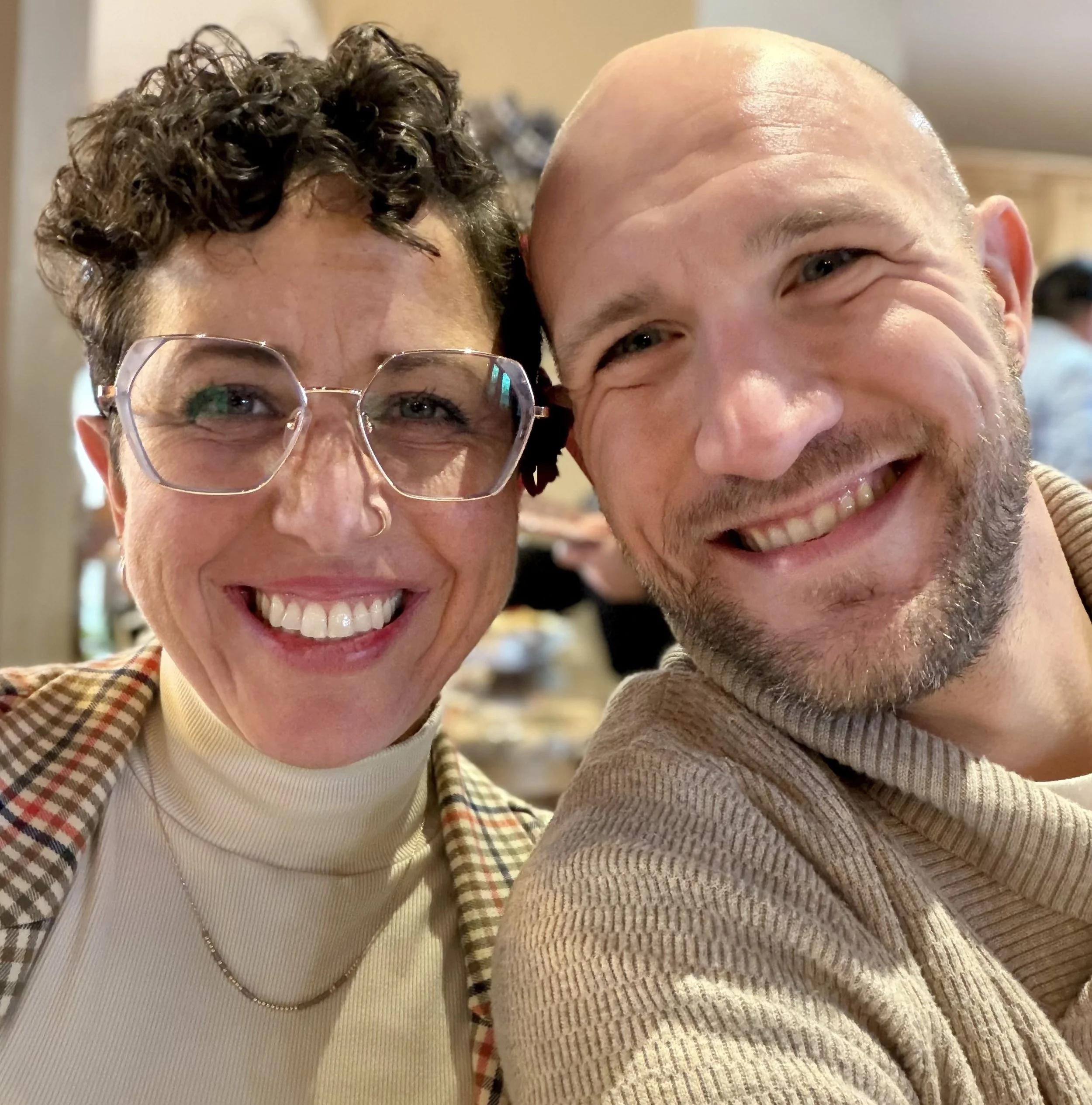 A woman with short curly hair, glasses, and a nose ring, smiling next to a man with a beard, bald head, and a big smile, in a warm indoor setting.