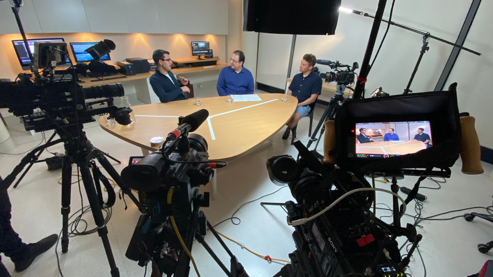 Three men sit around a table in a recording studio or TV set, surrounded by cameras and equipment, engaged in a discussion.