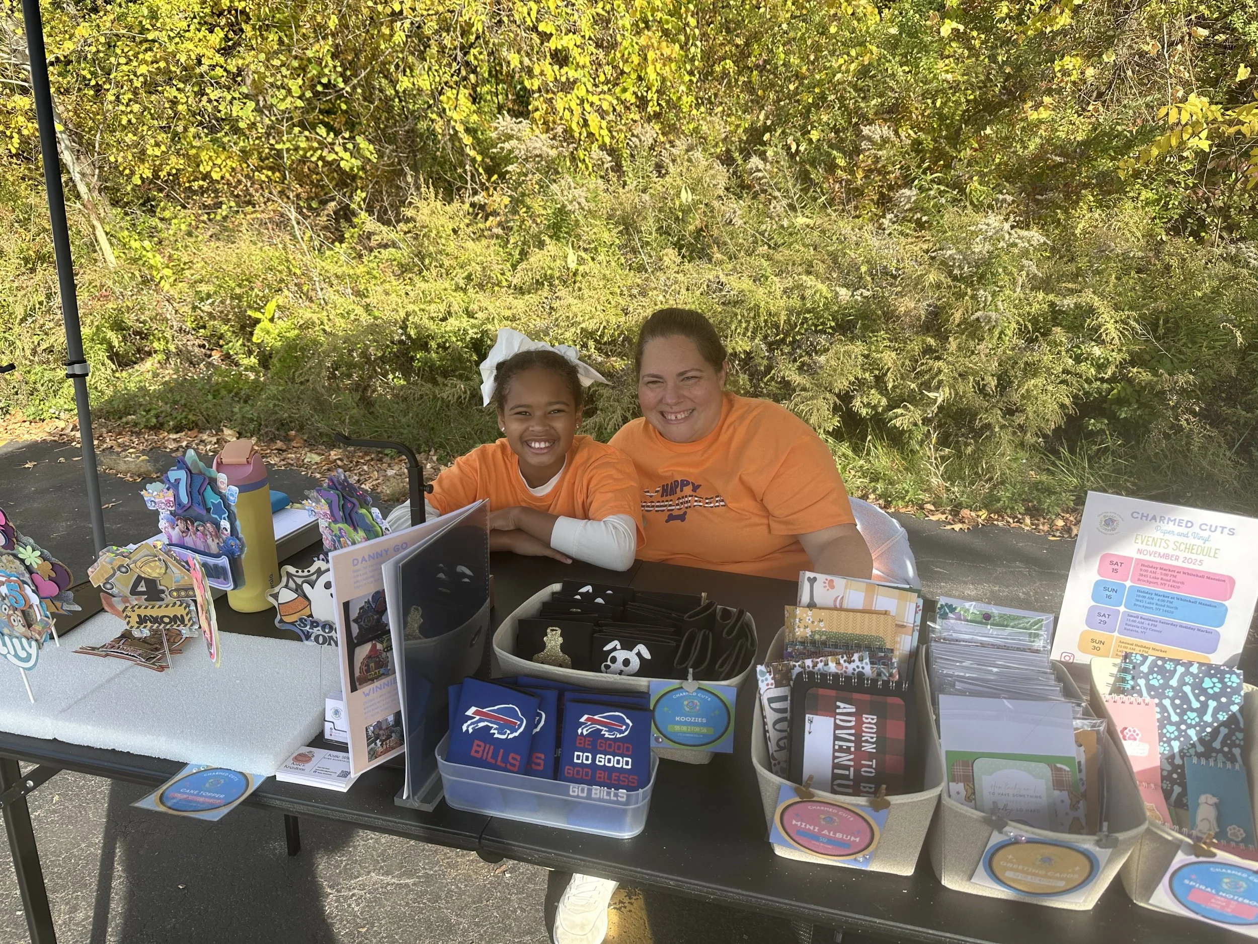 Two smiling girls sitting behind a table with various decorated items for sale, outdoors with trees in the background.