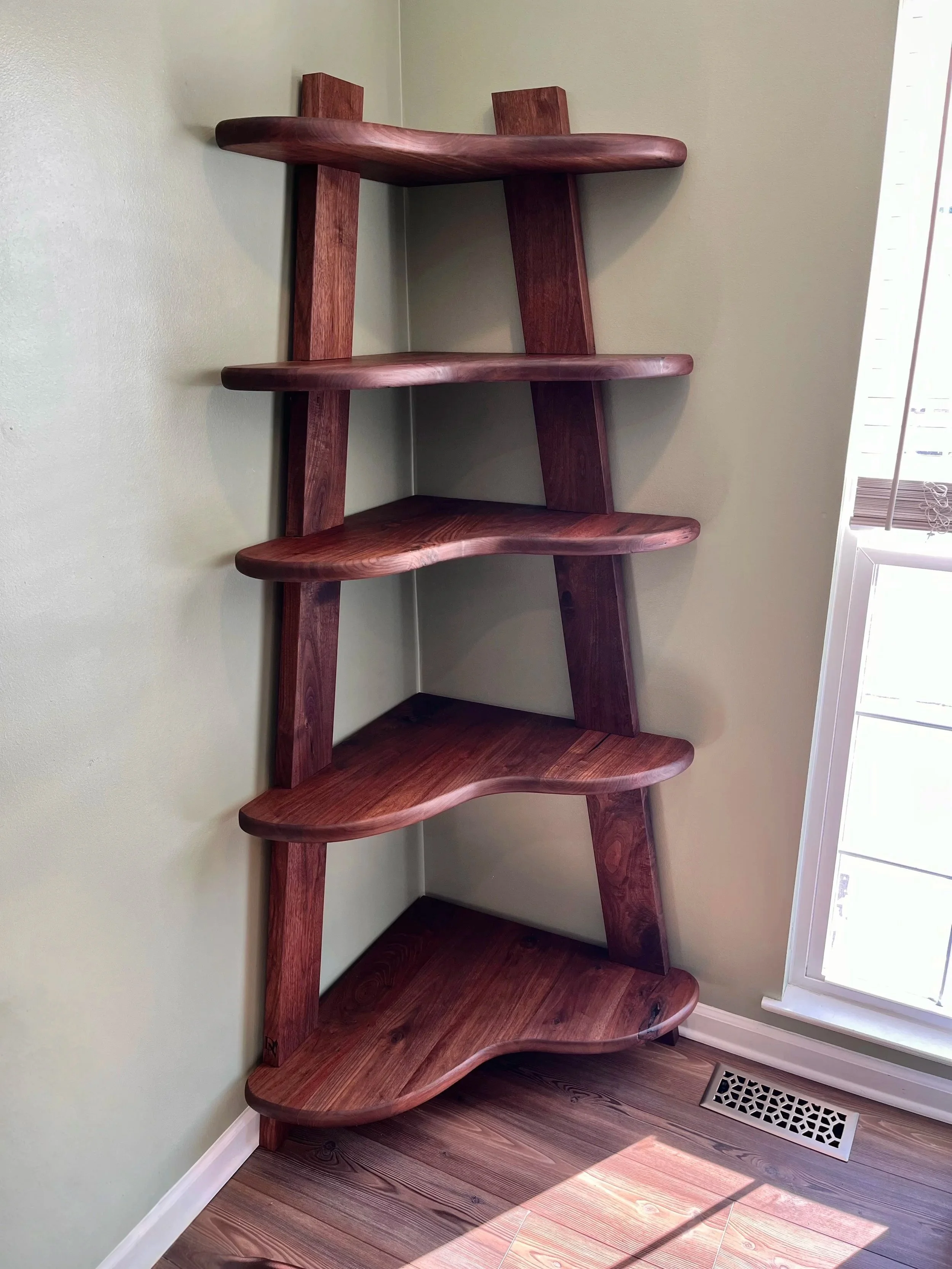 Wooden corner bookshelf with five curved shelves, placed in a corner near a window and a floor vent.