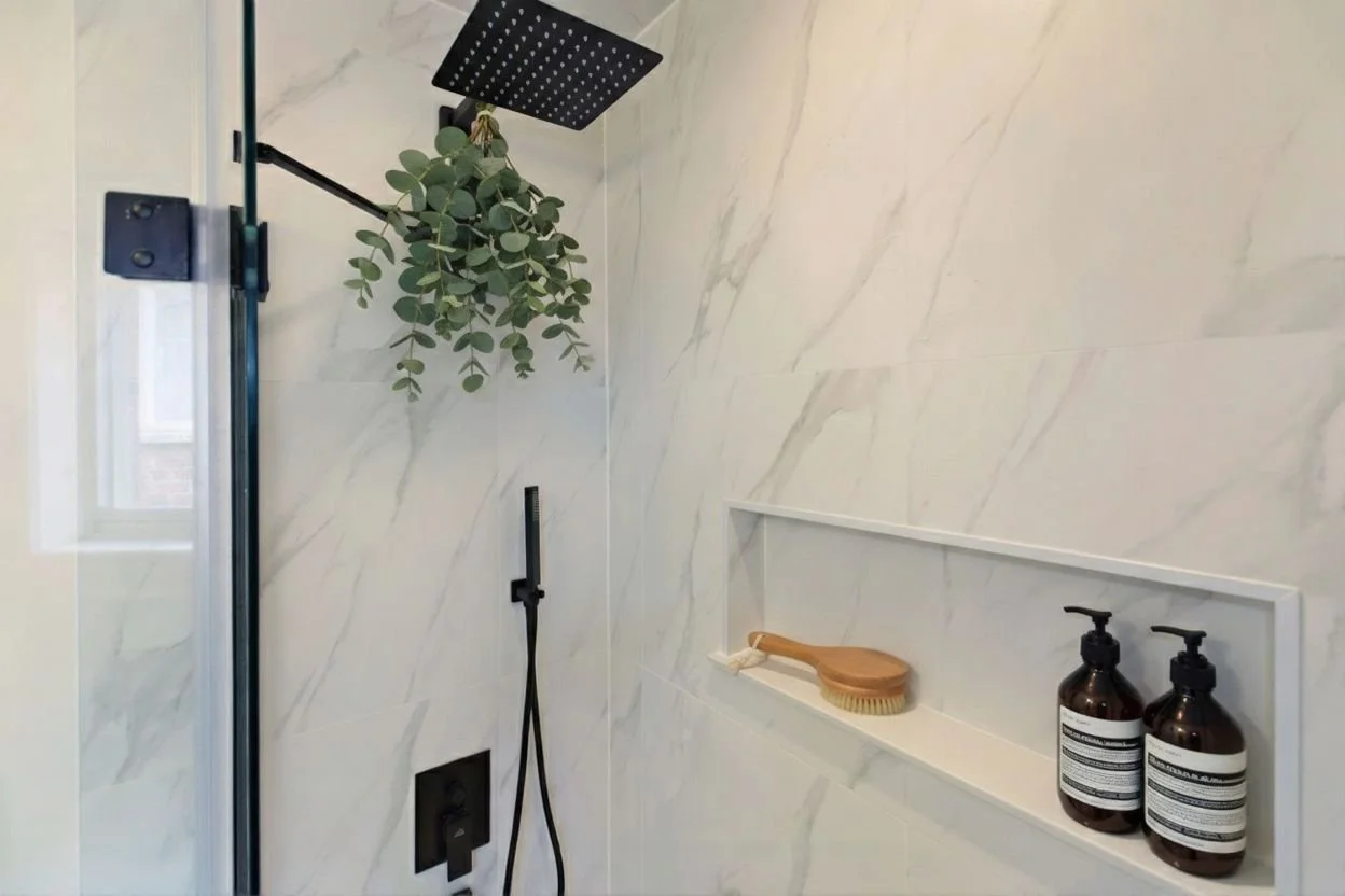 Bathroom shower with marble walls, a rain shower head, a shower wand, a speaker, and a niche containing two bottles and a scrub brush, with a hanging plant.