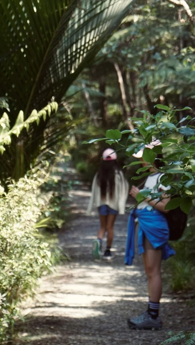 Two people walking along a forest trail surrounded by greenery, with one person in the foreground and the other in the background, both wearing casual outdoor clothing.