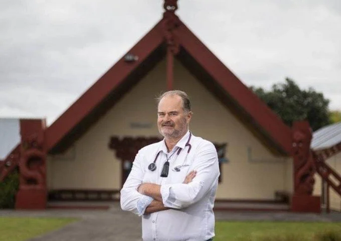 A male doctor standing outside in front of a traditional Polynesian-style building, with arms crossed and smiling.