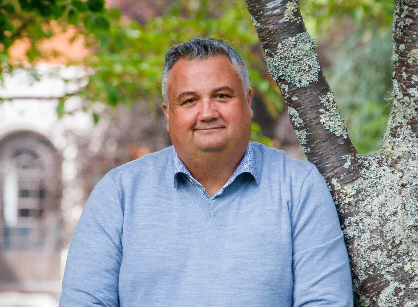 A middle-aged man with short gray hair leaning against a tree outdoors. He is wearing a light blue sweater over a collared shirt, and he is smiling. The background shows blurred trees and a building.