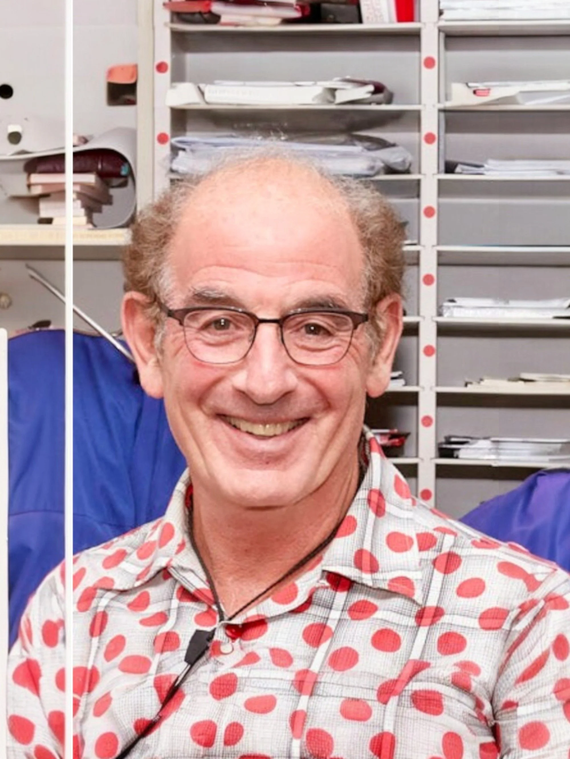 Smiling man with glasses, wearing a polka dot shirt, in front of a filing cabinet filled with papers.