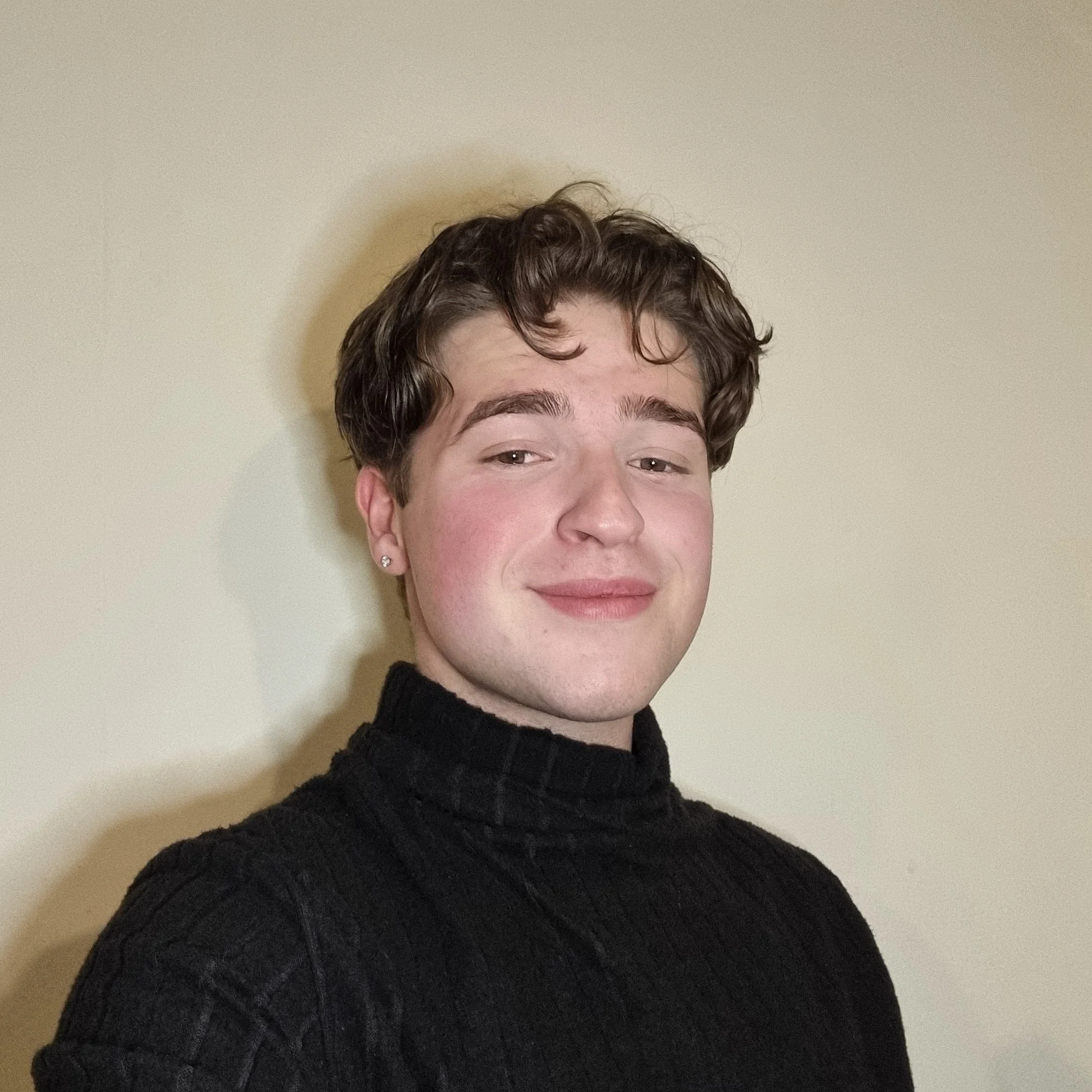 A young man with short curly brown hair, wearing a black turtleneck sweater, smiling slightly against a plain beige wall.