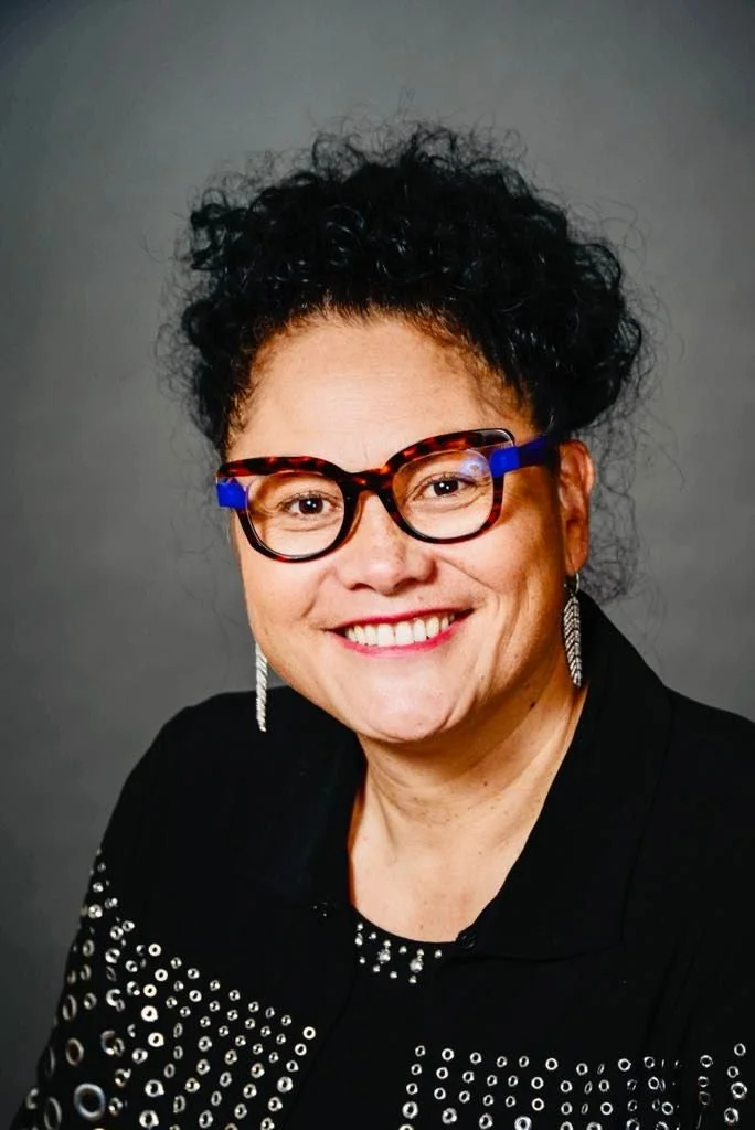 A woman with curly black hair, wearing tortoiseshell glasses with blue accents, silver earrings, and a black top with silver embellishments, smiling against a gray background.