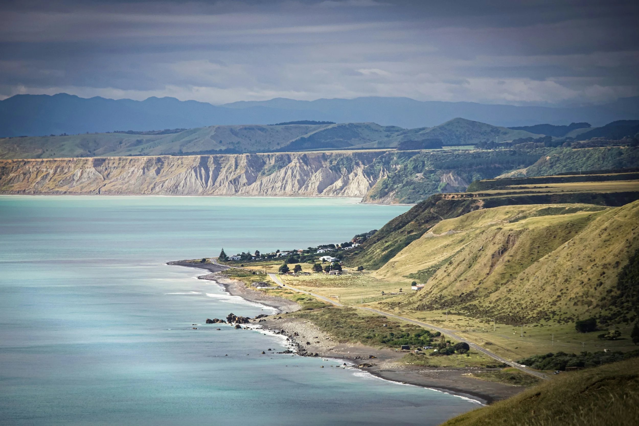 Coastal landscape with rolling green hills, a shoreline, and calm blue ocean under a cloudy sky, featuring distant mountains.