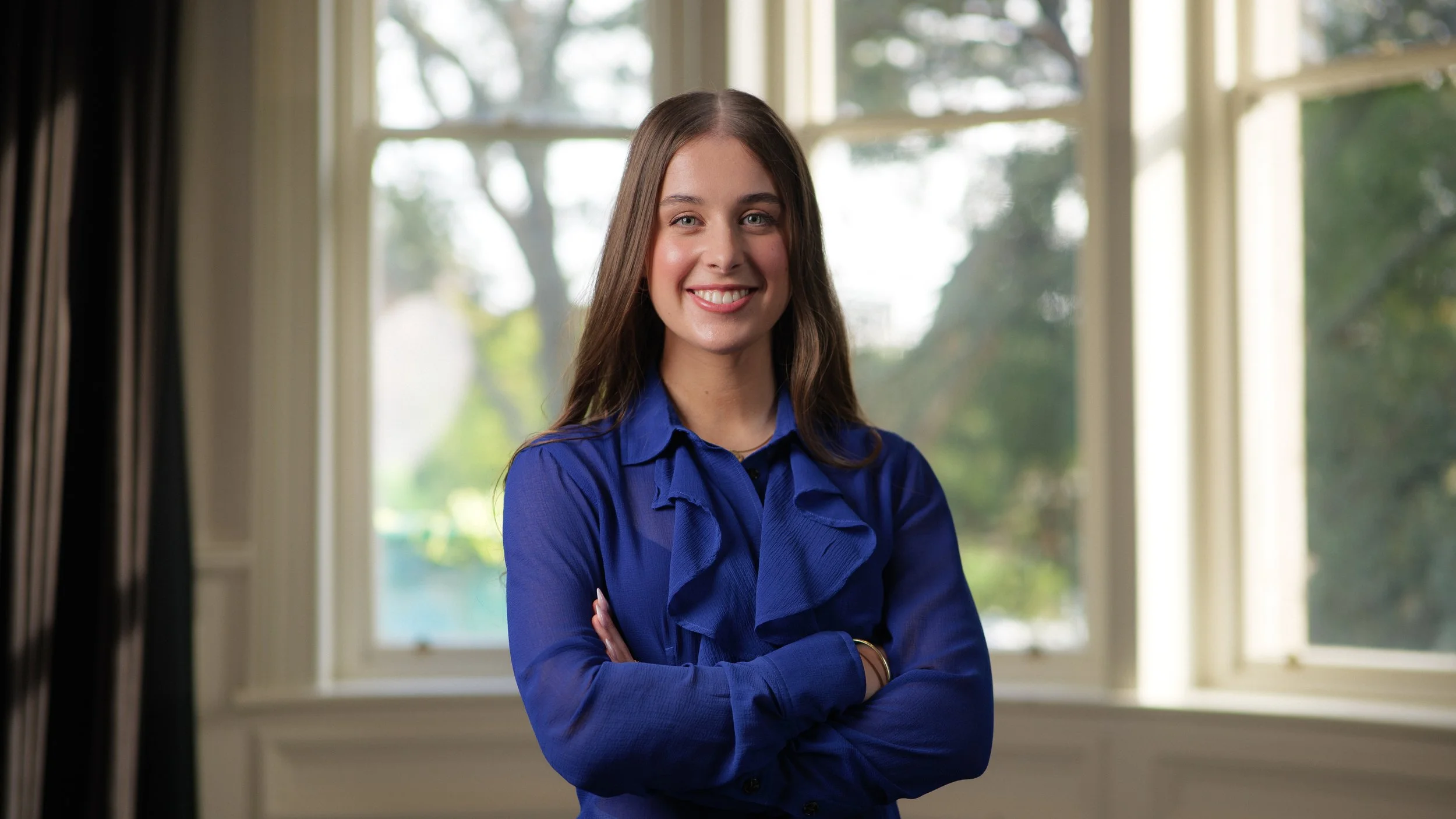 A young woman with long brown hair wearing a blue blouse, standing in a room with large windows and greenery outside, smiling with arms crossed.