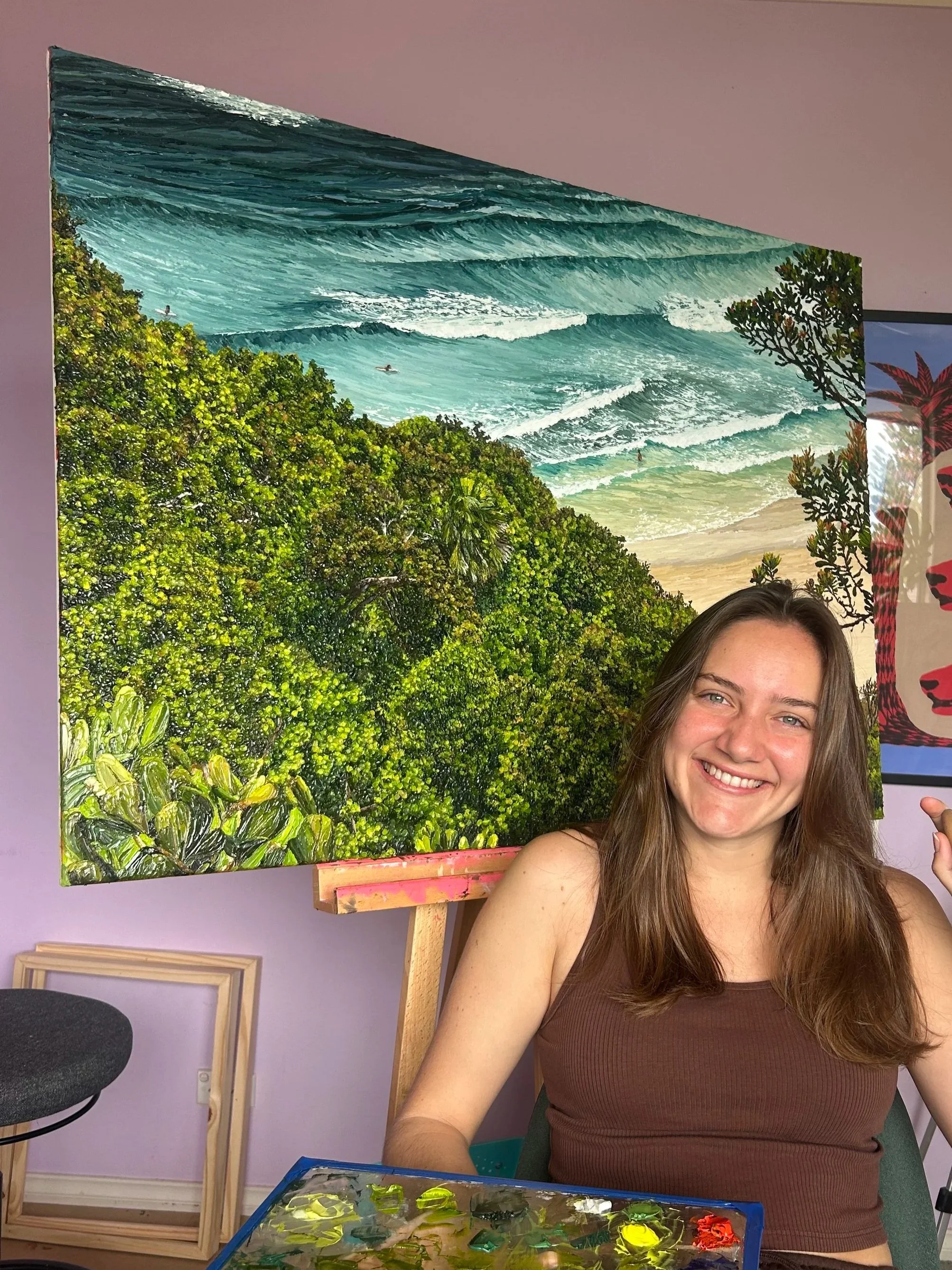 Young woman with long brown hair smiling and sitting in front of a vibrant painting of the ocean with waves and greenery.