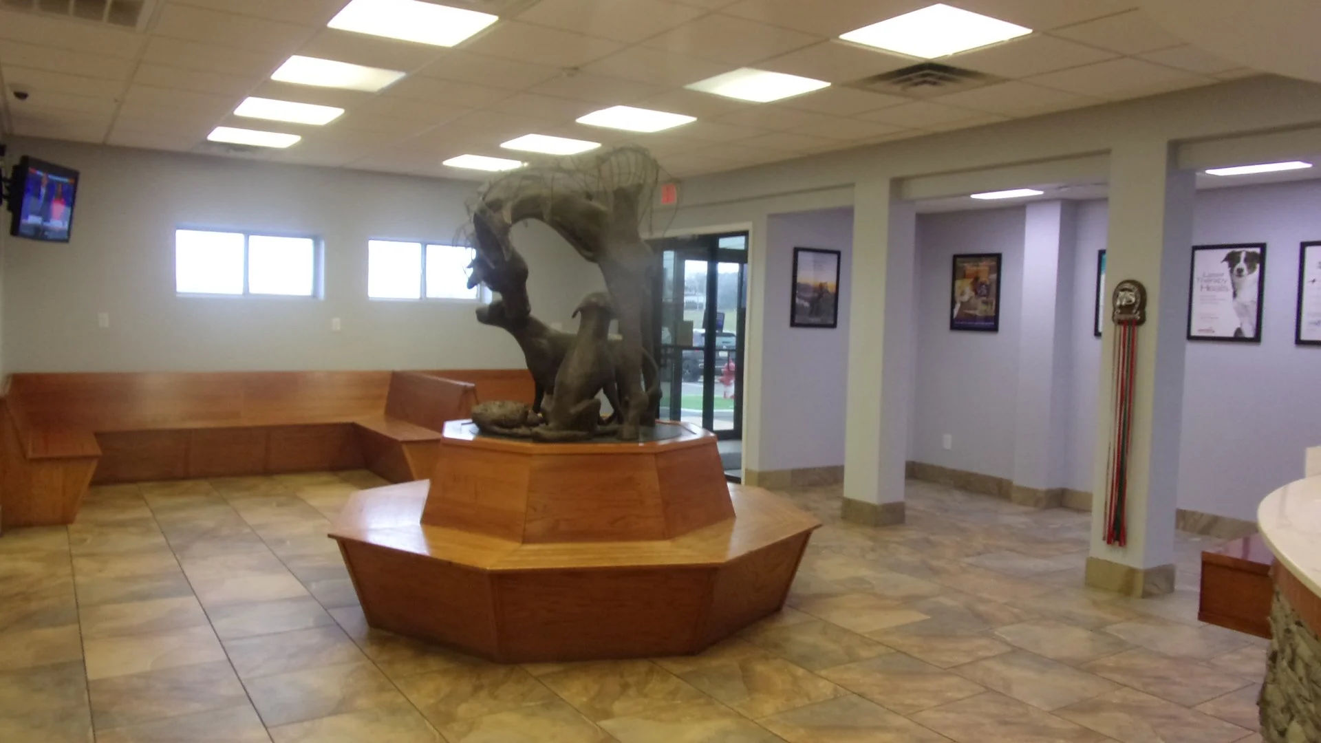 Empty lobby with wooden benches, a central sculpture of a dragon, framed posters on walls, and a wall-mounted television.