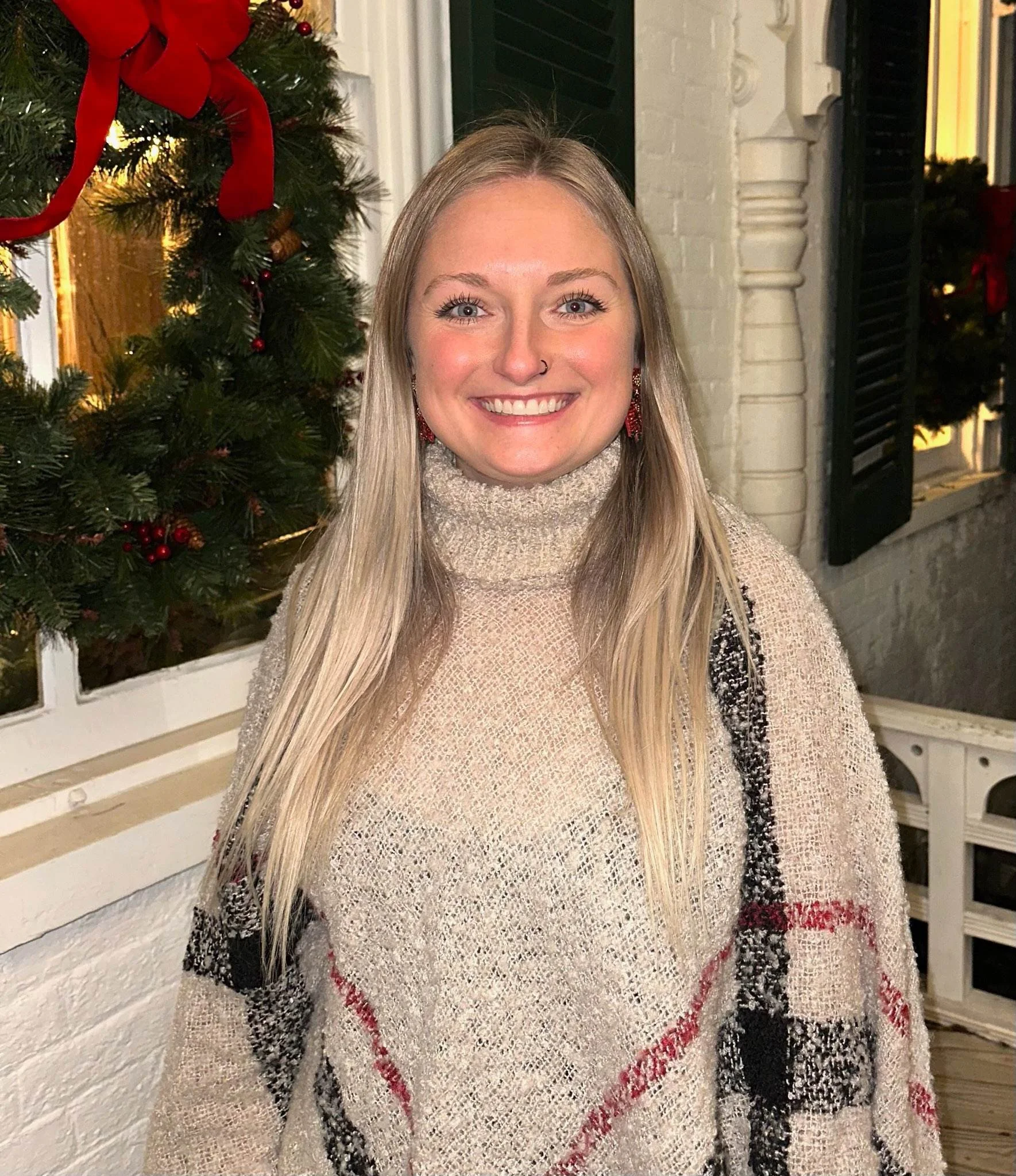 A young woman with long blonde hair, smiling, wearing a beige turtleneck sweater and red earrings, standing outside a house decorated with a Christmas tree and holiday lights.