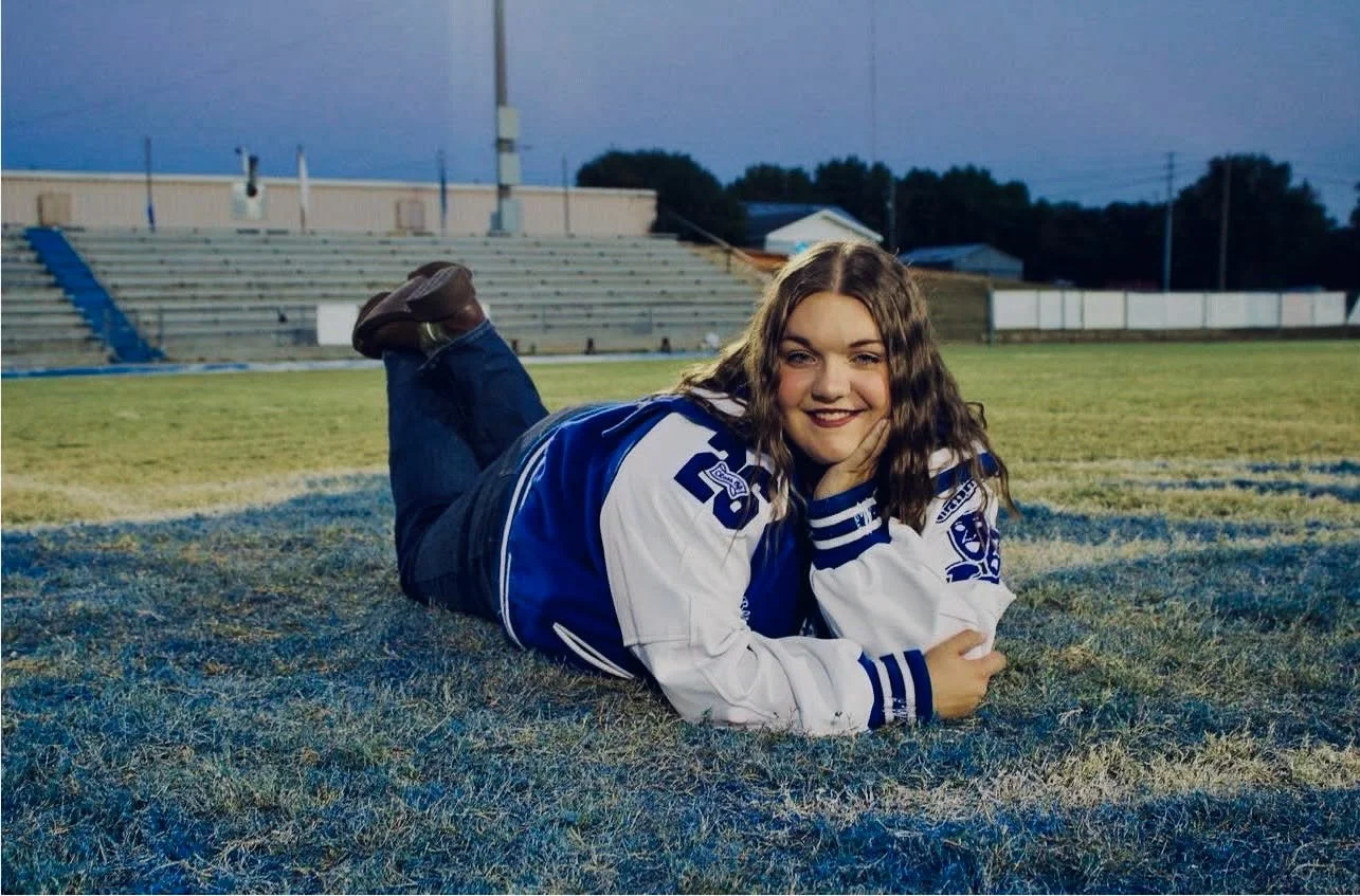 A young woman lies on her stomach on a football field at dusk, smiling at the camera. She is wearing a blue and white football jersey and jeans.