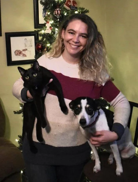 A woman holding a black cat in one arm and a black and white dog in the other, standing in a living room with a decorated Christmas tree in the background.