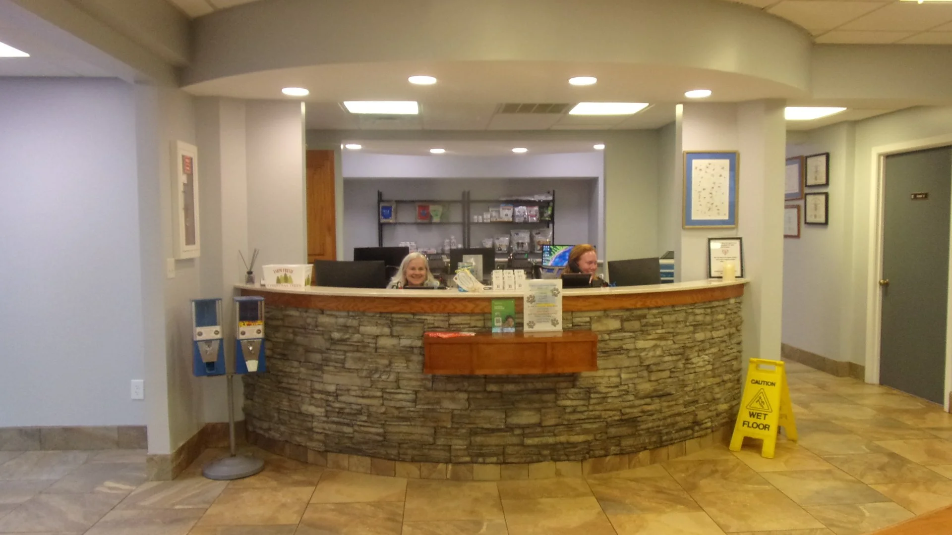 Front desk with two smiling women, one with long gray hair and the other with blonde hair, behind a curved stone counter inside a building, with framed pictures on the wall and a yellow wet floor caution sign on the floor.